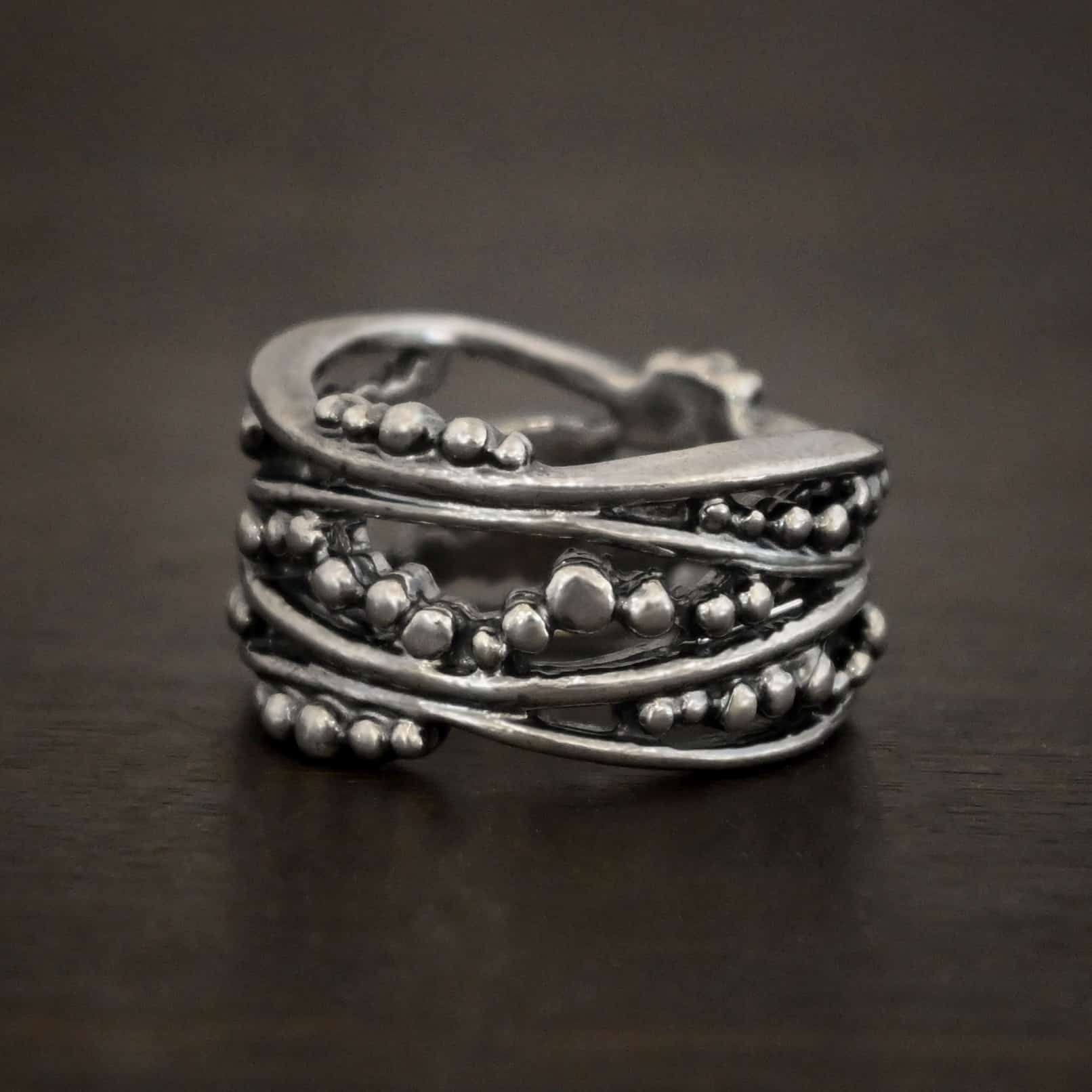 River Ring Photo of the "River" Ring. Made out of sterling silver. It has multiple lines of silver that intercept at random places, but all of them meet at the back to create the band of the ring. There are silver dots scattered all over the ring in between the gaps of where the silver does not meet. The top and bottom of the lines flow and create an uneven ring band. Ring is sitting against a dark background.