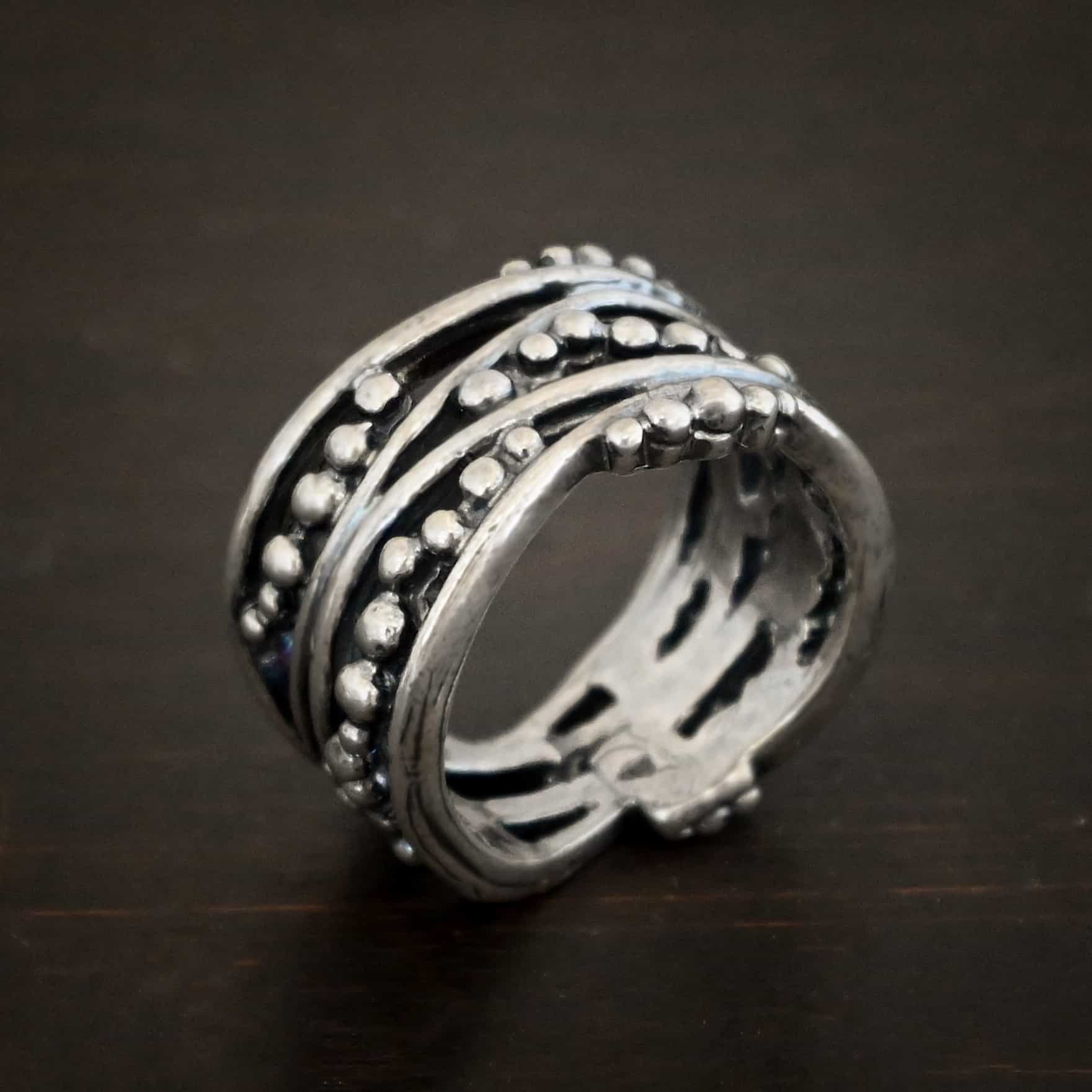 River Ring Photo of the "River" Ring. Made out of sterling silver. It has multiple lines of silver that intercept at random places, but all of them meet at the back to create the band of the ring. There are silver dots scattered all over the ring in between the gaps of where the silver does not meet. The top and bottom of the lines flow and create an uneven ring band. Ring is sitting against a dark background.