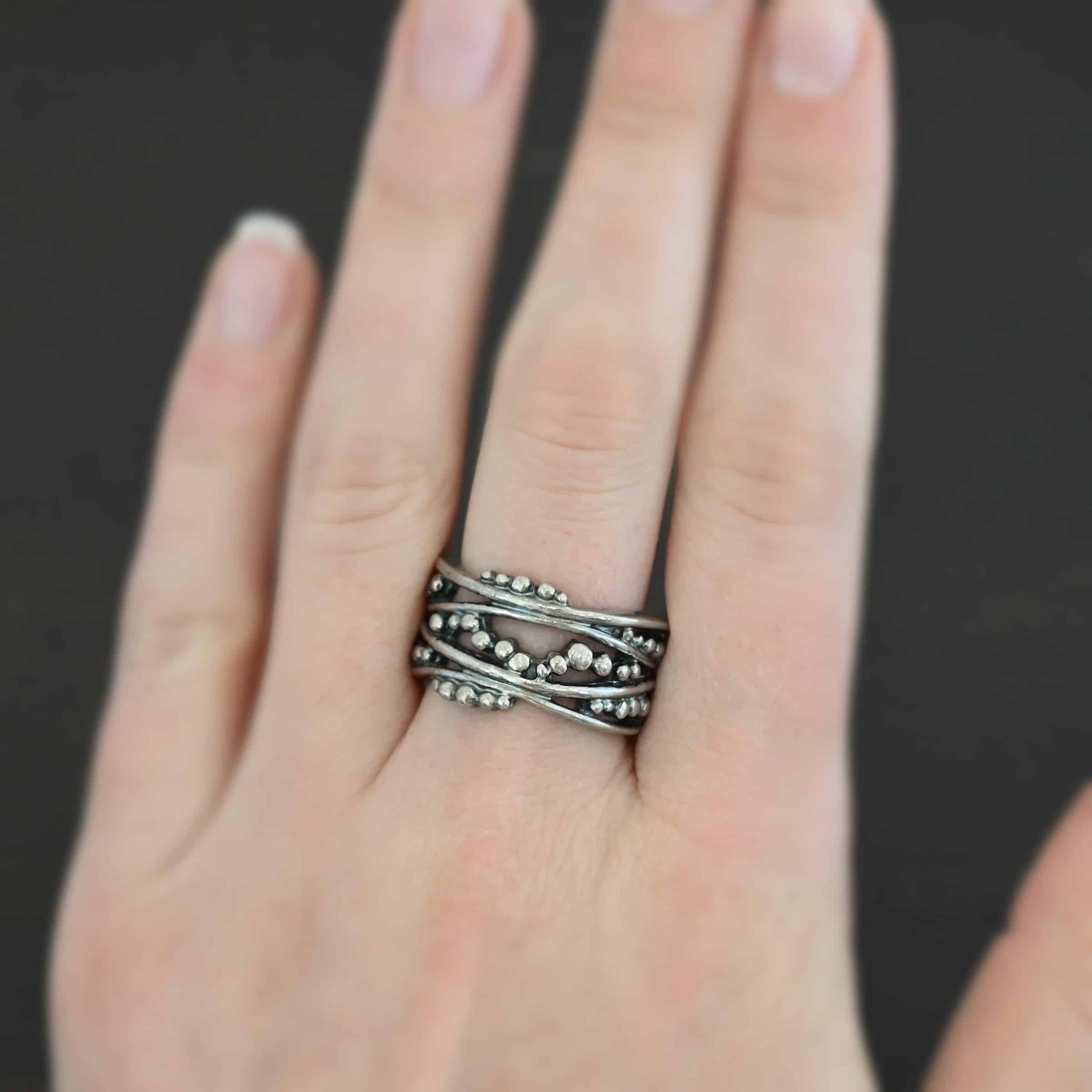 River Ring Photo of the "River" Ring. Made out of sterling silver. It has multiple lines of silver that intercept at random places, but all of them meet at the back to create the band of the ring. There are silver dots scattered all over the ring in between the gaps of where the silver does not meet. The top and bottom of the lines flow and create an uneven ring band. Ring is worn by a model to demonstrate size.