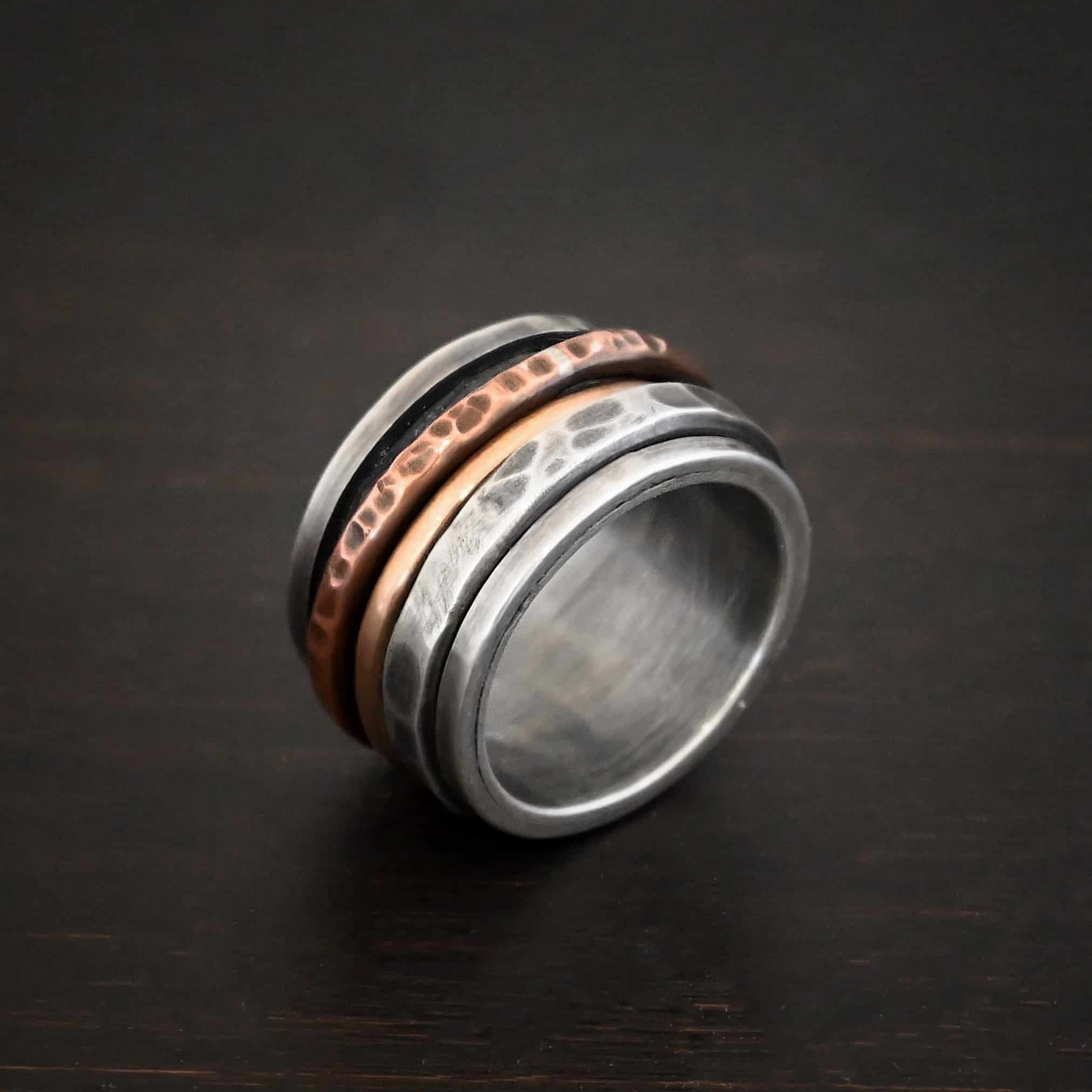 Royal Revolver Ring Photo of the "Royal Revolver" Ring. Made out of sterling silver, copper, and 14k gold. The top and bottom of the ring are silver, and there is a gap in the center of the ring. The gap is taken up by a hammered copper spinner, a smooth 14k gold spinner, and a thick, hammered sterling silver spinner. Each band in the center spins around. Ring is sitting against a dark background.