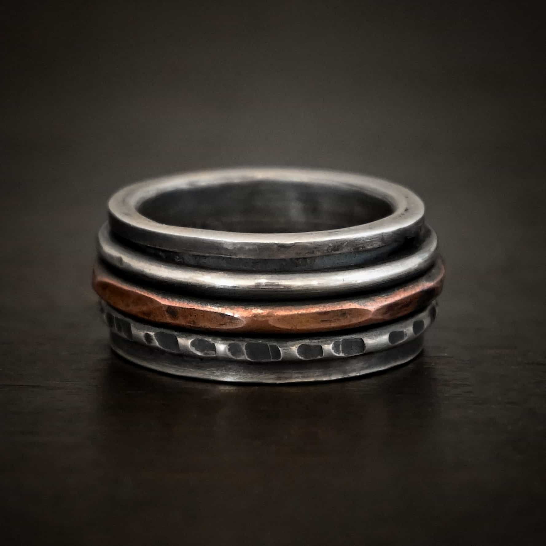 Revolver Ring Photo of the "Revolver" Ring. Made out of sterling silver and copper. The top and bottom of the ring are silver, and there is a gap in the center of the ring. The gap is taken up by a hammered copper spinner and a thick, hammered sterling silver spinner, as well as a smooth sterling silver spinner. Each band in the center spins around. Ring is sitting against a dark background.