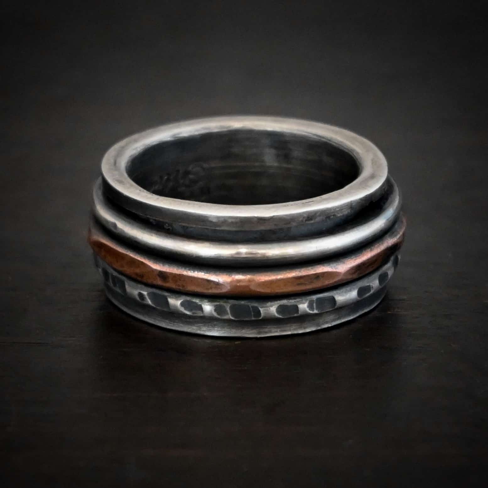Revolver Ring Photo of the "Revolver" Ring. Made out of sterling silver and copper. The top and bottom of the ring are silver, and there is a gap in the center of the ring. The gap is taken up by a hammered copper spinner and a thick, hammered sterling silver spinner, as well as a smooth sterling silver spinner. Each band in the center spins around. Ring is sitting against a dark background.