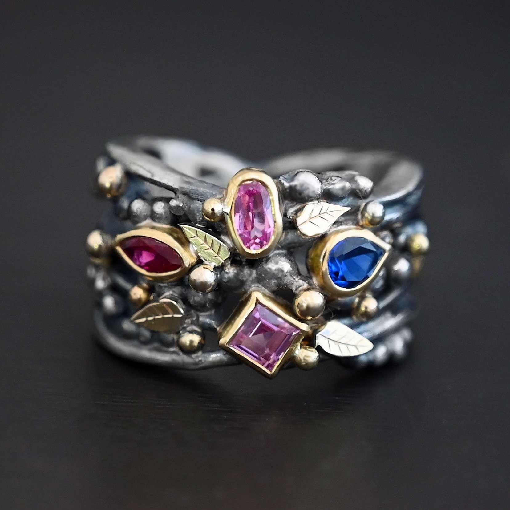Mothers Birthstone Ring Photo of a custom ring made for a mother with birthstones. There top stone is a pink zirconia that is oval shaped. The bottom stone is also a pink zirconia, but diamond shaped. The left side stone is a marquise shaped ruby. The right side stone is a teardrop shaped sapphire. The ring is a mix of silver and gold. The band is silver, but all of the stones are set in gold and there are gold balls scattered all around the ring. Ring is sitting against a dark background.
