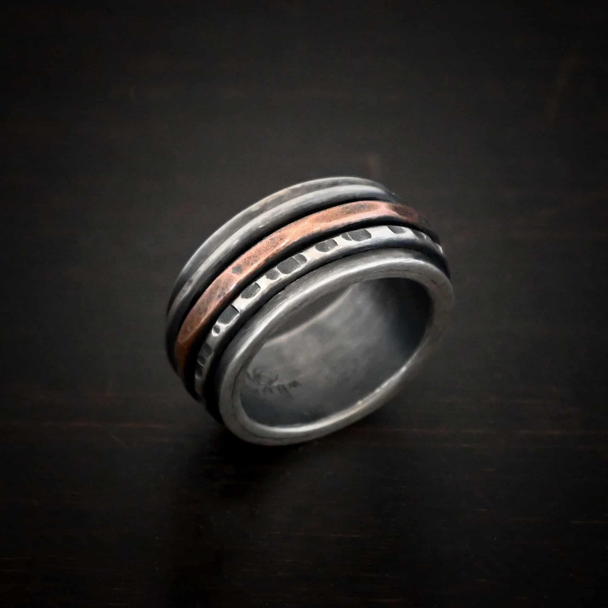 Revolver Ring Photo of the "Revolver" Ring. Made out of sterling silver and copper. The top and bottom of the ring are silver, and there is a gap in the center of the ring. The gap is taken up by a hammered copper spinner and a thick, hammered sterling silver spinner, as well as a smooth sterling silver spinner. Each band in the center spins around. Ring is sitting against a dark background.