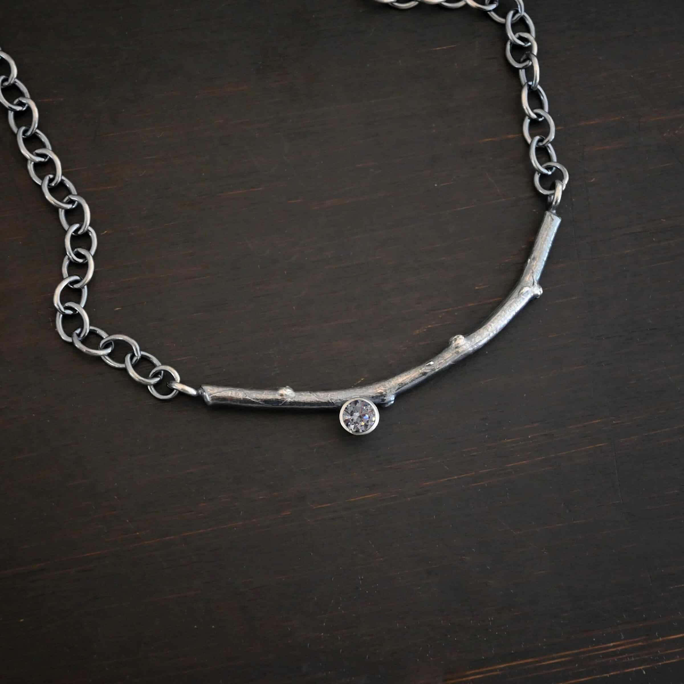 Frosted Twig Necklace Photo of the "Frosted Twig" Necklace. Made out of a casted sterling silver twig with small bumps around it. The twig is slightly curved. On the left side of the curve, soldered to the bottom, is a medium sized circular cubic zirconia that is set in silver. On both ends of the twig there are two loops that are connected to a chain that connects at the back to wear the necklace. Necklace is lying against a dark background.