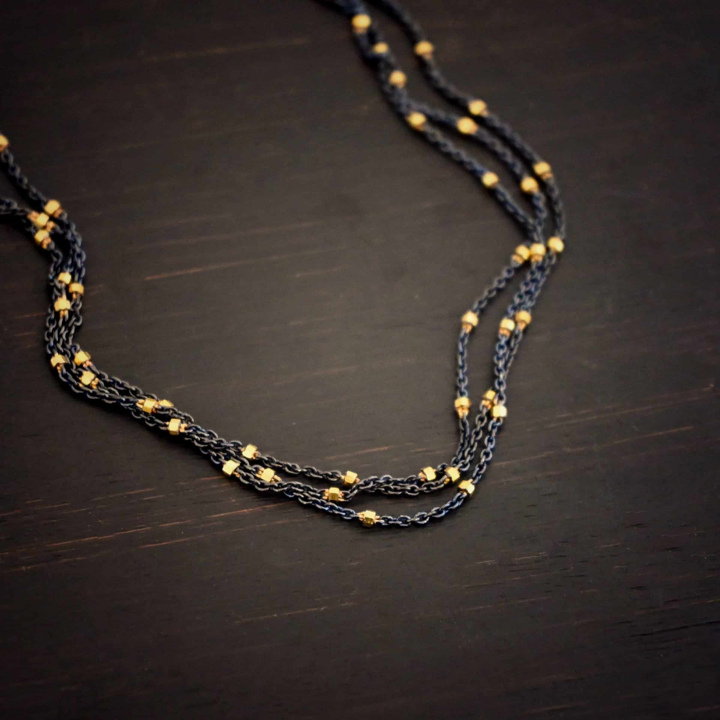 Night in Paris Chain "Night In Paris" Chain as shown up close. Sterling Silver & 14k yellow gold filled beads that are 1.5mm thick. The chain is 1.2mm thick. The chain is made out of three separate strands that meet at the back to make one chain. Chain is shown against a dark background.