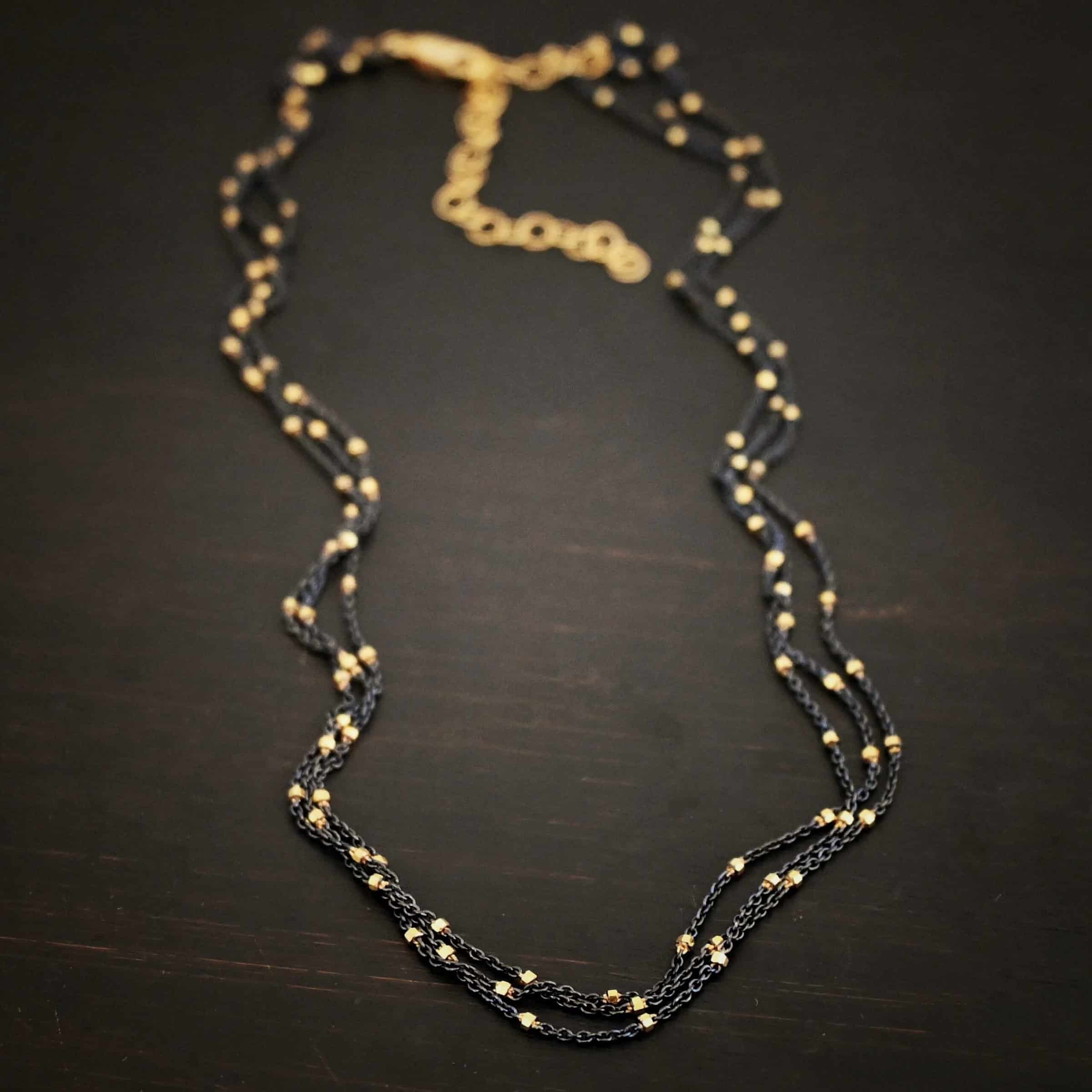 Night in Paris Chain "Night In Paris" Chain as shown up close. Sterling Silver & 14k yellow gold filled beads that are 1.5mm thick. The chain is 1.2mm thick. The chain is made out of three separate strands that meet at the back to make one chain. Chain is shown against a dark background.