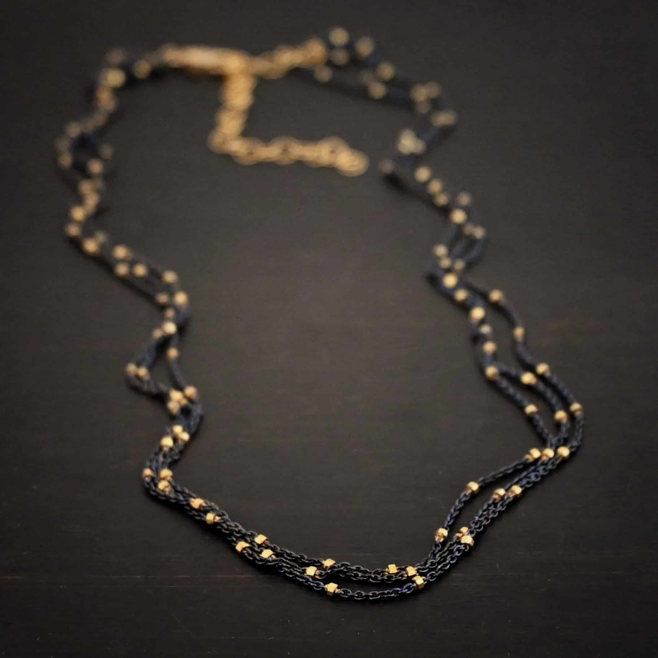 Night in Paris Chain "Night In Paris" Chain as shown up close. Sterling Silver & 14k yellow gold filled beads that are 1.5mm thick. The chain is 1.2mm thick. The chain is made out of three separate strands that meet at the back to make one chain. Chain is shown against a dark background.