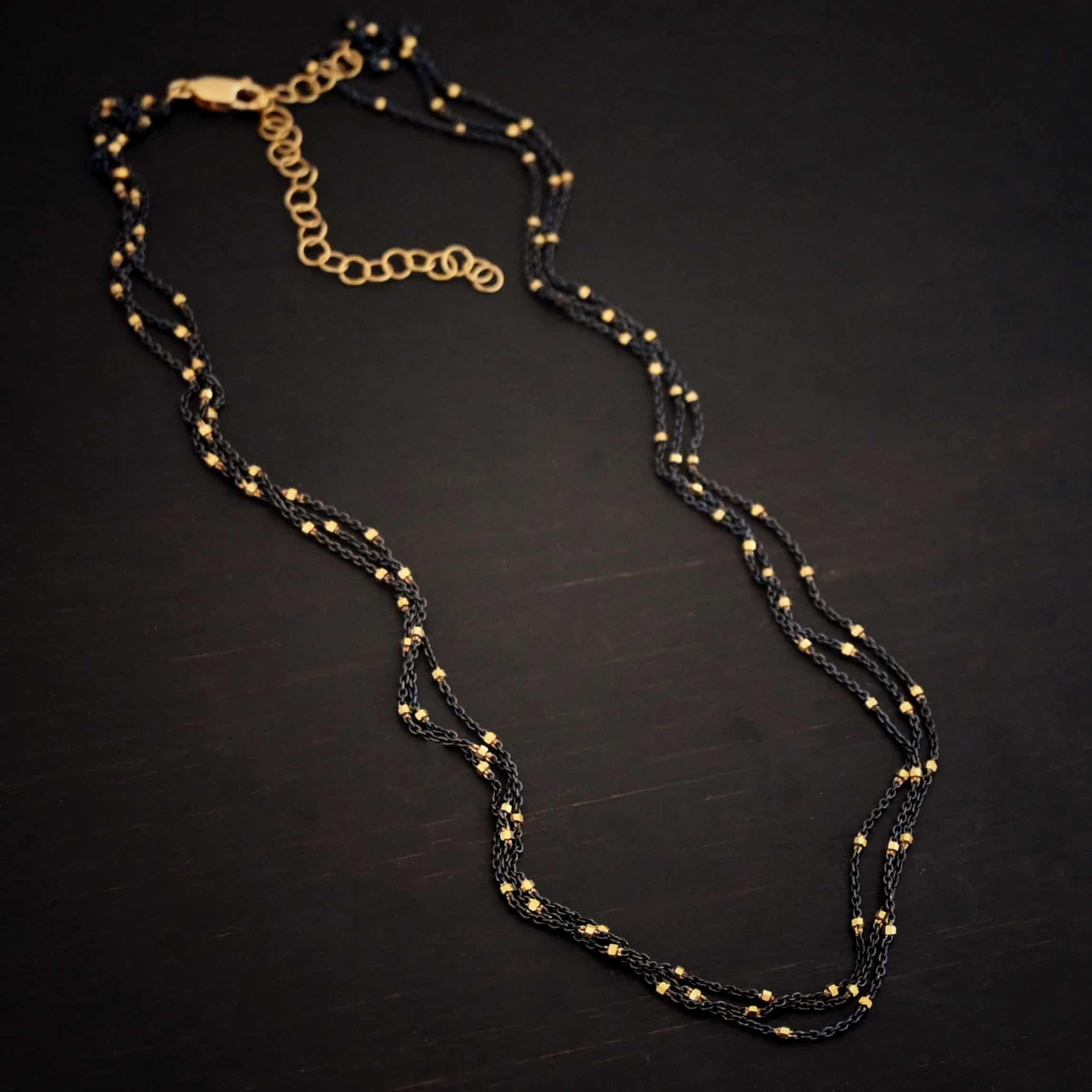 Night in Paris Chain "Night In Paris" Chain as shown up close. Sterling Silver & 14k yellow gold filled beads that are 1.5mm thick. The chain is 1.2mm thick. The chain is made out of three separate strands that meet at the back to make one chain. Chain is shown against a dark background.