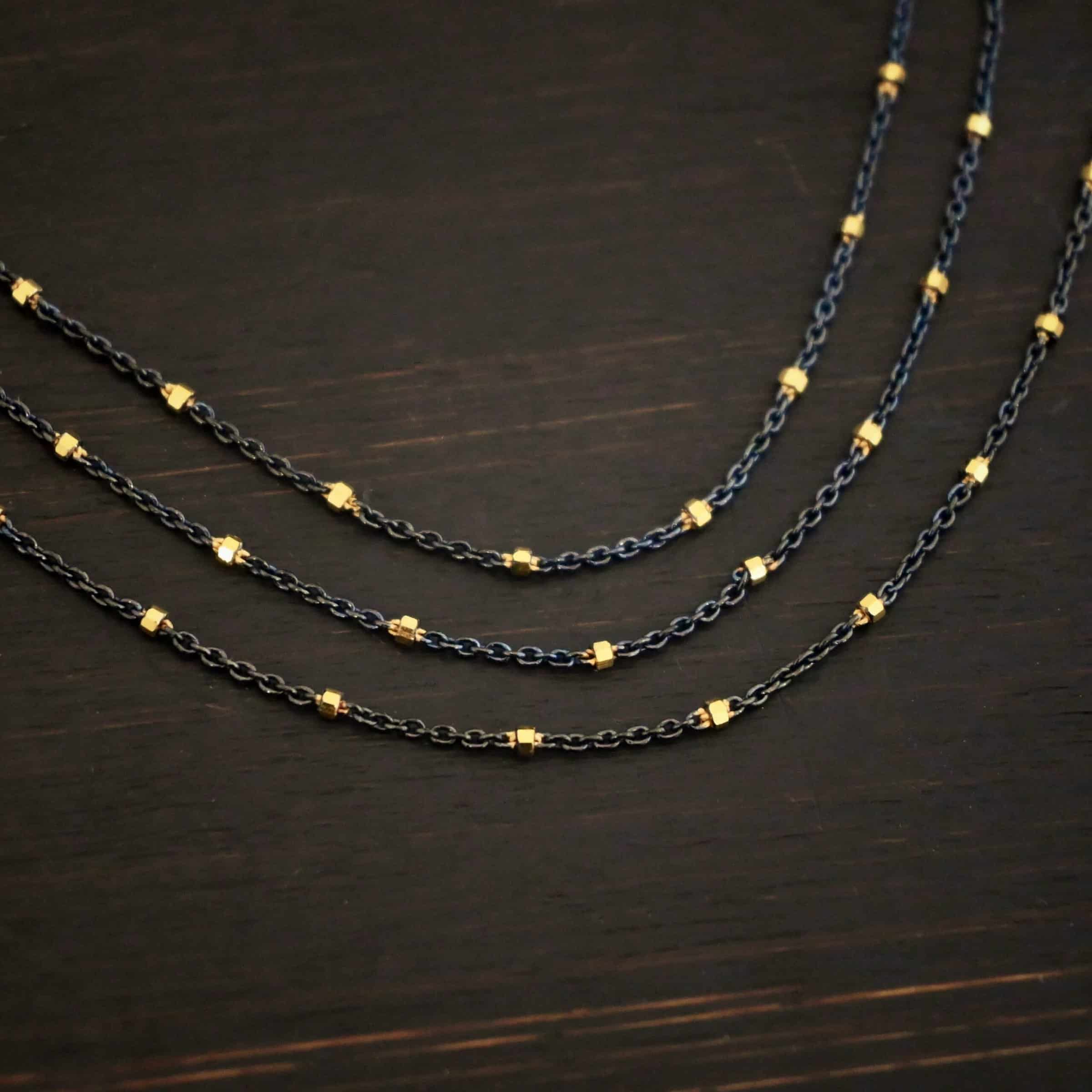 Night in Paris Chain "Night In Paris" Chain as shown up close. Sterling Silver & 14k yellow gold filled beads that are 1.5mm thick. The chain is 1.2mm thick. The chain is made out of three separate strands that meet at the back to make one chain. Chain is shown against a dark background.