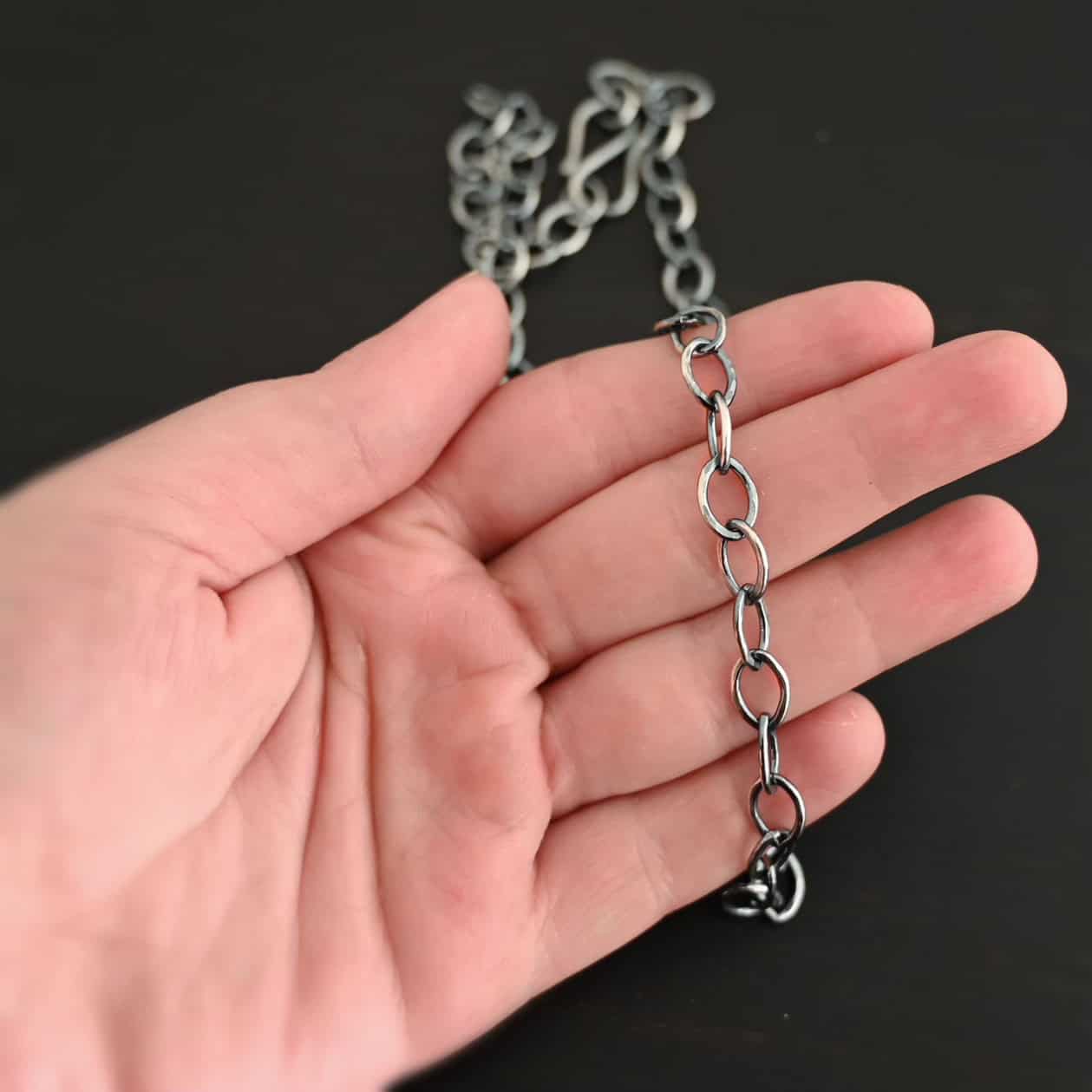 Sky Chain, Hammered, Oxidized Oxidized sterling silver Sky Chain that is made with large oval links that are hammered and are all connected together laying on a hand that is resting against a dark background