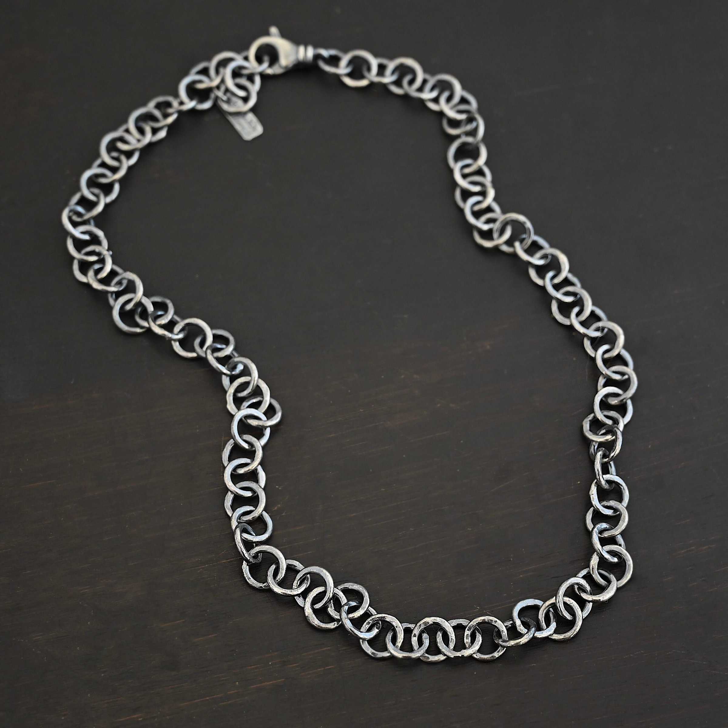 Close up of the "Wyatt" Chain. Chain is handmade out of sterling silver and has thick circle links that are hammered and oxidized. Chain has a lobster claw clasp and a rectangle tag that says "Wild Prairie Silver" on it. Chain is laying against a dark background.
