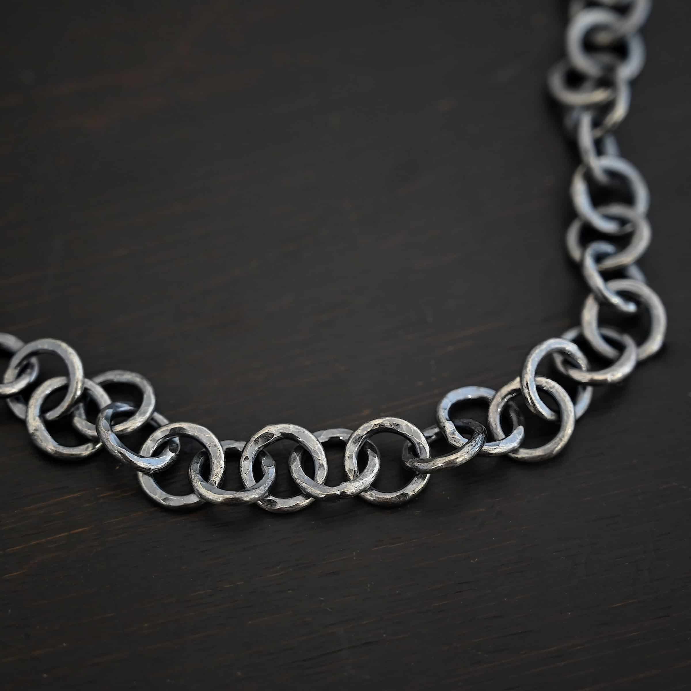 Close up of the "Wyatt" Chain. Chain is handmade out of sterling silver and has thick circle links that are hammered and oxidized. Chain is laying against a dark background.