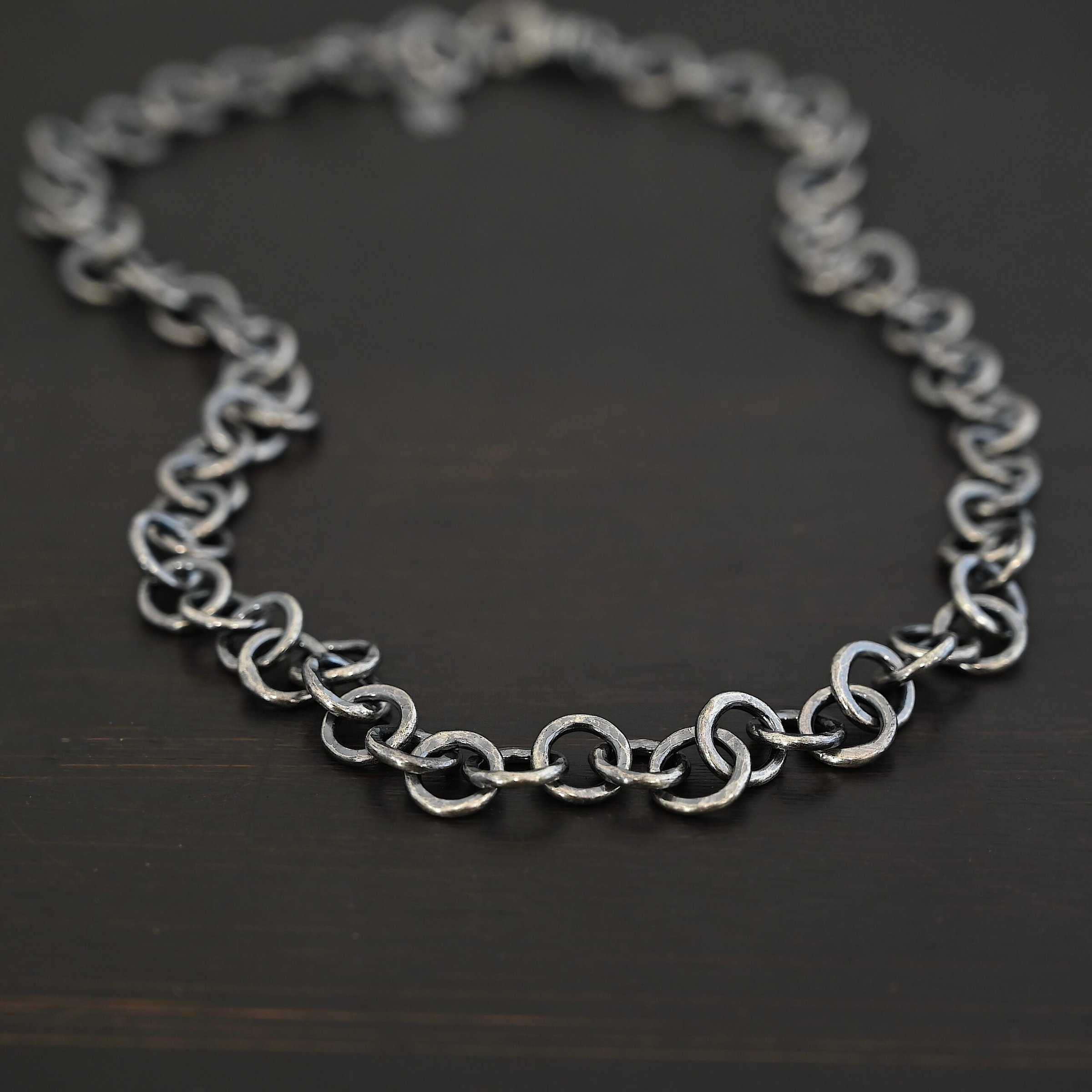 Close up of the "Wyatt" Chain. Chain is handmade out of sterling silver and has thick circle links that are hammered and oxidized. Chain is laying against a dark background.