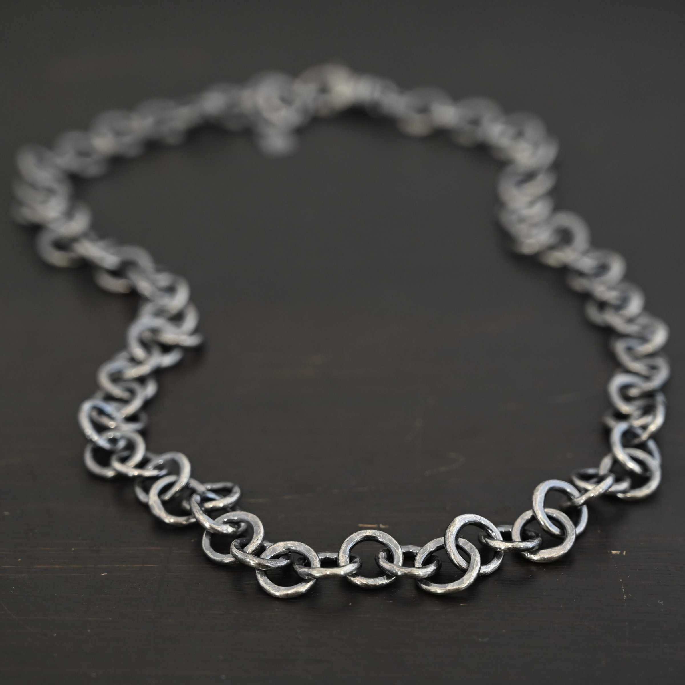 Close up of the "Wyatt" Chain. Chain is handmade out of sterling silver and has thick circle links that are hammered and oxidized. Chain is laying against a dark background.