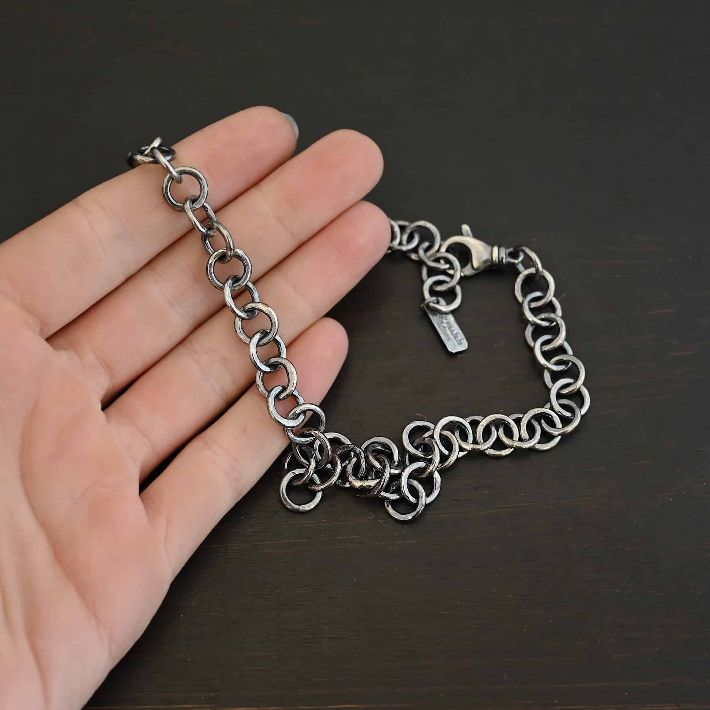 Close up of the "Wyatt" Chain. Chain is handmade out of sterling silver and has thick circle links that are hammered and oxidized. Chain has a lobster claw clasp and a rectangle tag that says "Wild Prairie Silver" on it. Chain is laying in the fingertips of a hand to demonstrate size.