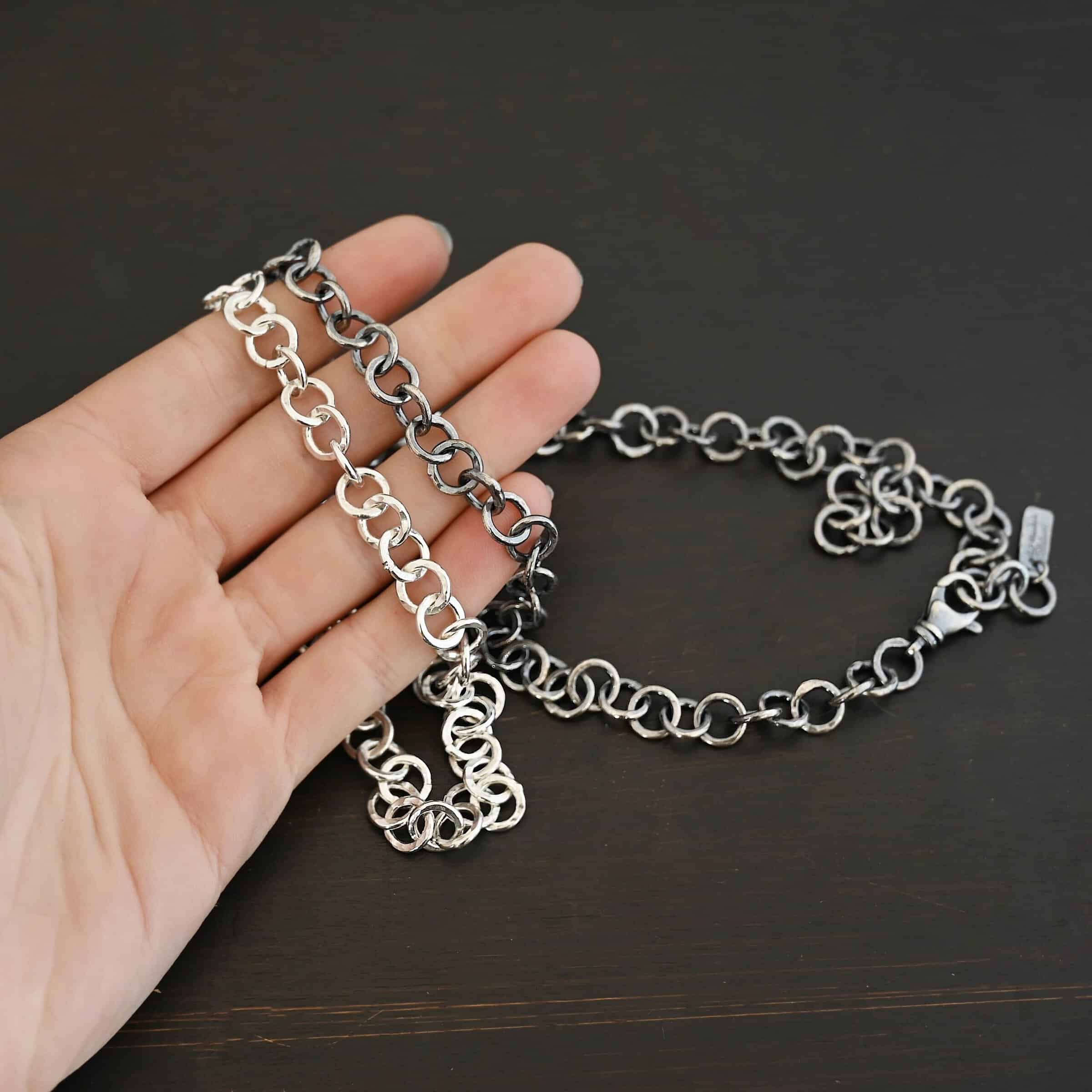 Close up of the "Wyatt" Chain. Chain is handmade out of sterling silver and has thick circle links that are hammered and shiny and oxidized. Chain has a lobster claw clasp and a rectangle tag that says "Wild Prairie Silver" on it. Chain is laying in the fingertips of a hand to demonstrate size.