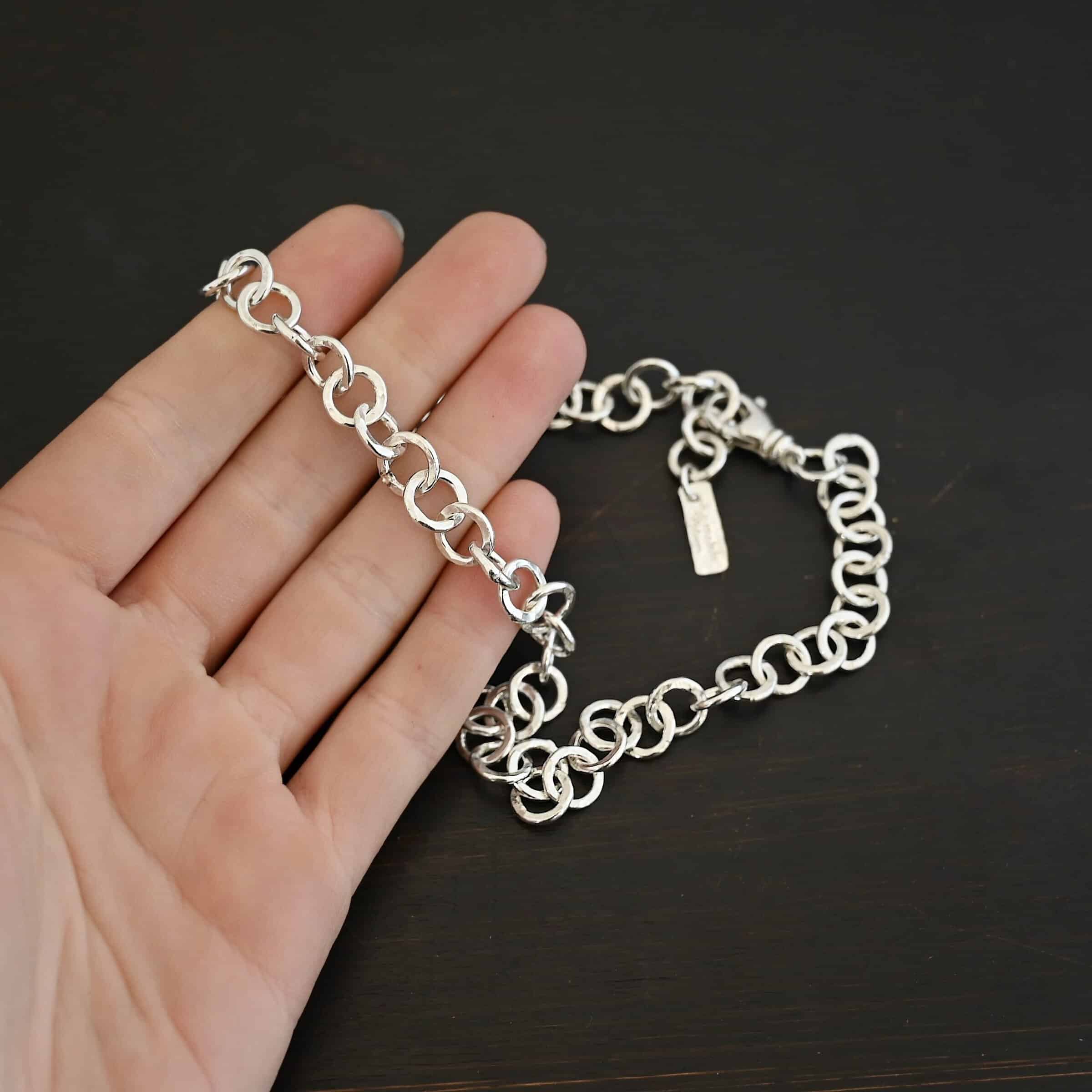 Close up of the "Wyatt" Chain. Chain is handmade out of sterling silver and has thick circle links that are hammered and shiny. chain has a lobster claw clasp and a rectangle tag that says "Wild Prairie Silver" on it. Chain is laying in the fingertips of a hand to demonstrate size.