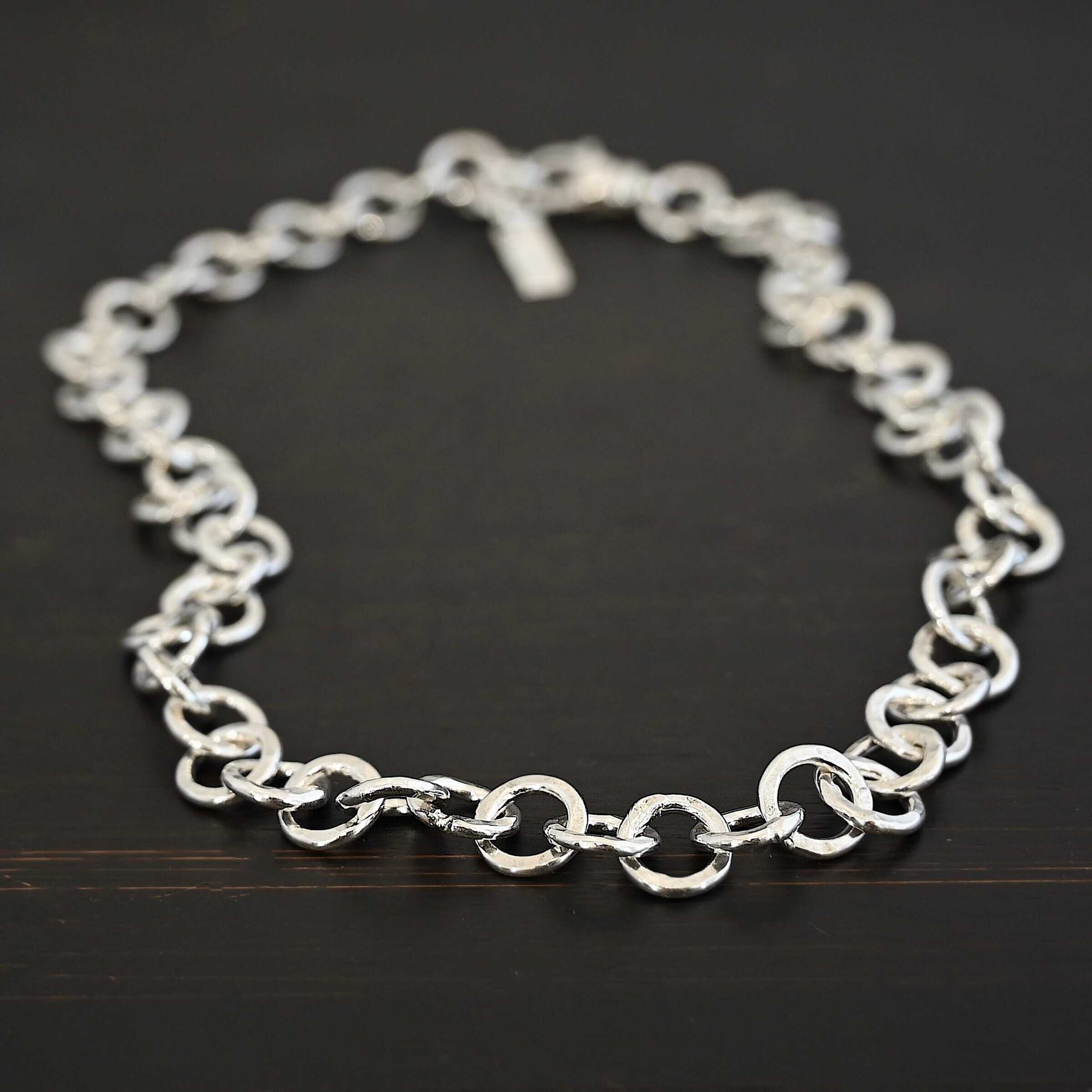 Close up of the "Wyatt" Chain. Chain is handmade out of sterling silver and has thick circle links that are hammered and shiny. chain has a lobster claw clasp and a rectangle tag that says "Wild Prairie Silver" on it. Chain is laying against a dark background.