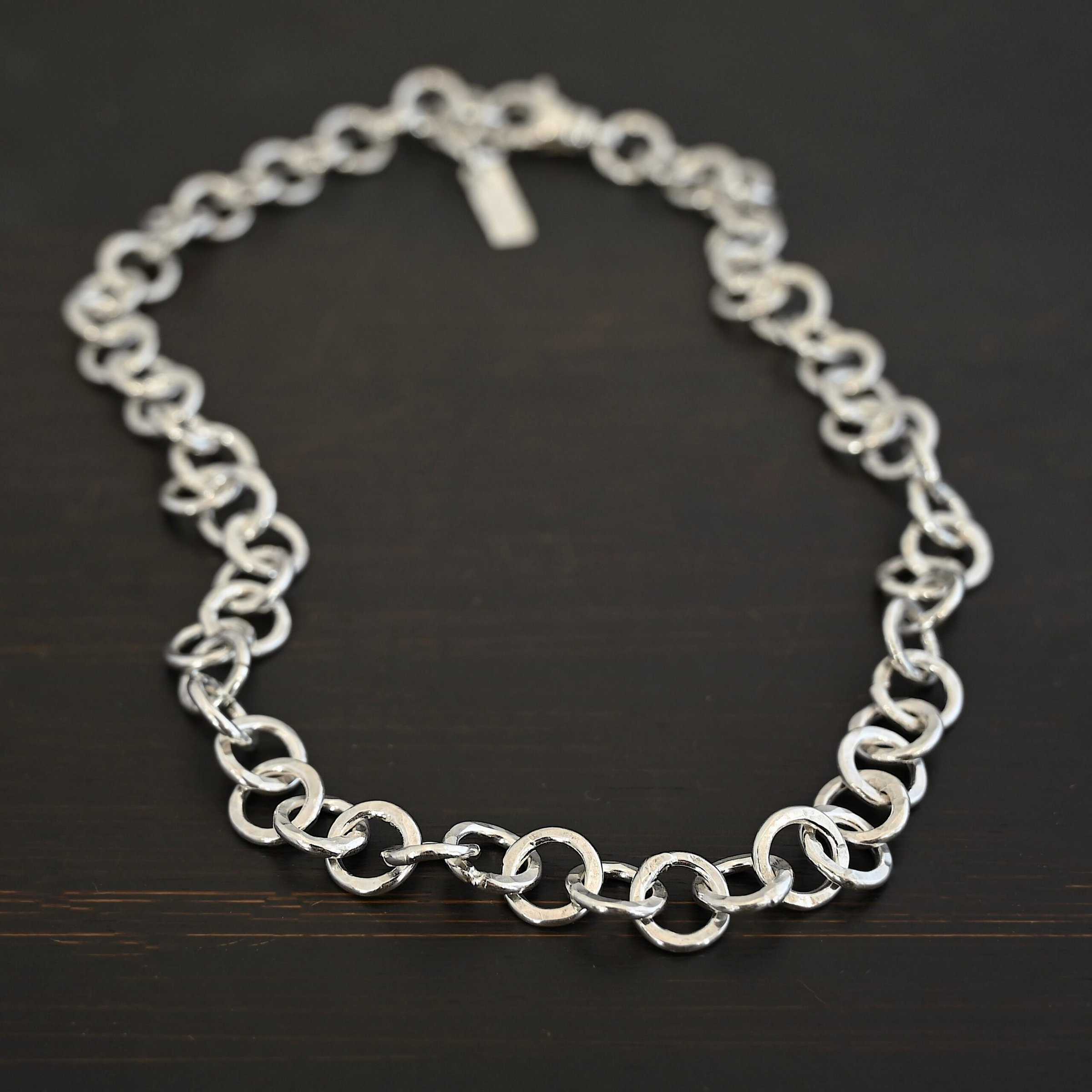 Close up of the "Wyatt" Chain. Chain is handmade out of sterling silver and has thick circle links that are hammered and shiny. chain has a lobster claw clasp and a rectangle tag that says "Wild Prairie Silver" on it. Chain is laying against a dark background.