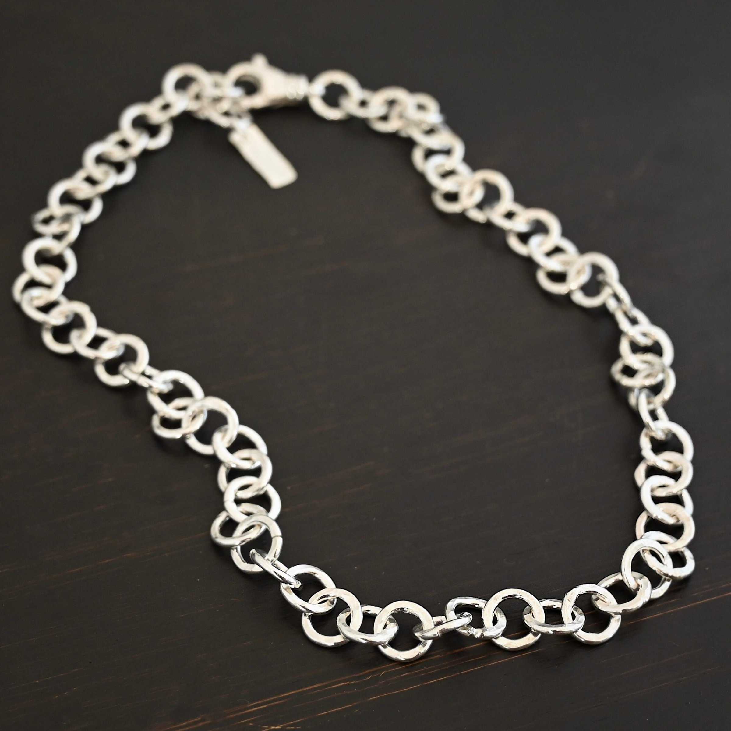 Close up of the "Wyatt" Chain. Chain is handmade out of sterling silver and has thick circle links that are hammered and shiny. chain has a lobster claw clasp and a rectangle tag that says "Wild Prairie Silver" on it. Chain is laying against a dark background.