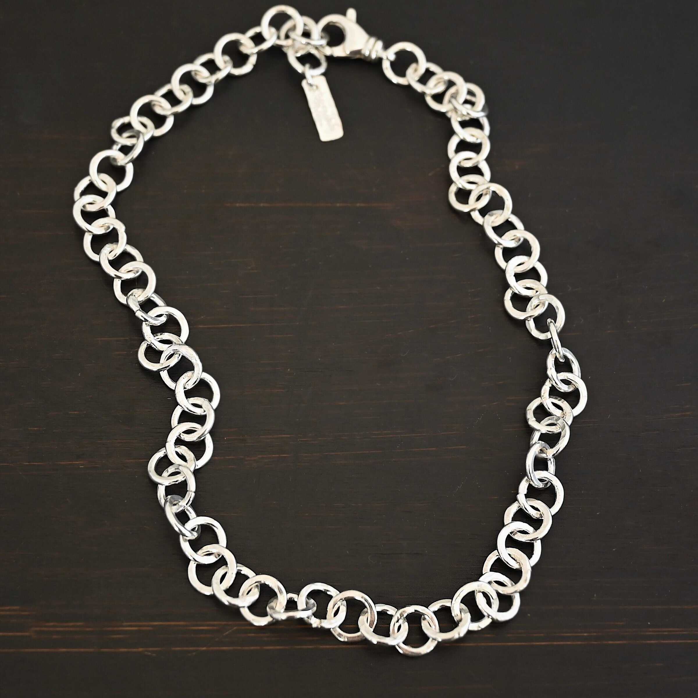 Close up of the "Wyatt" Chain. Chain is handmade out of sterling silver and has thick circle links that are hammered and shiny. chain has a lobster claw clasp and a rectangle tag that says "Wild Prairie Silver" on it. Chain is laying against a dark background.