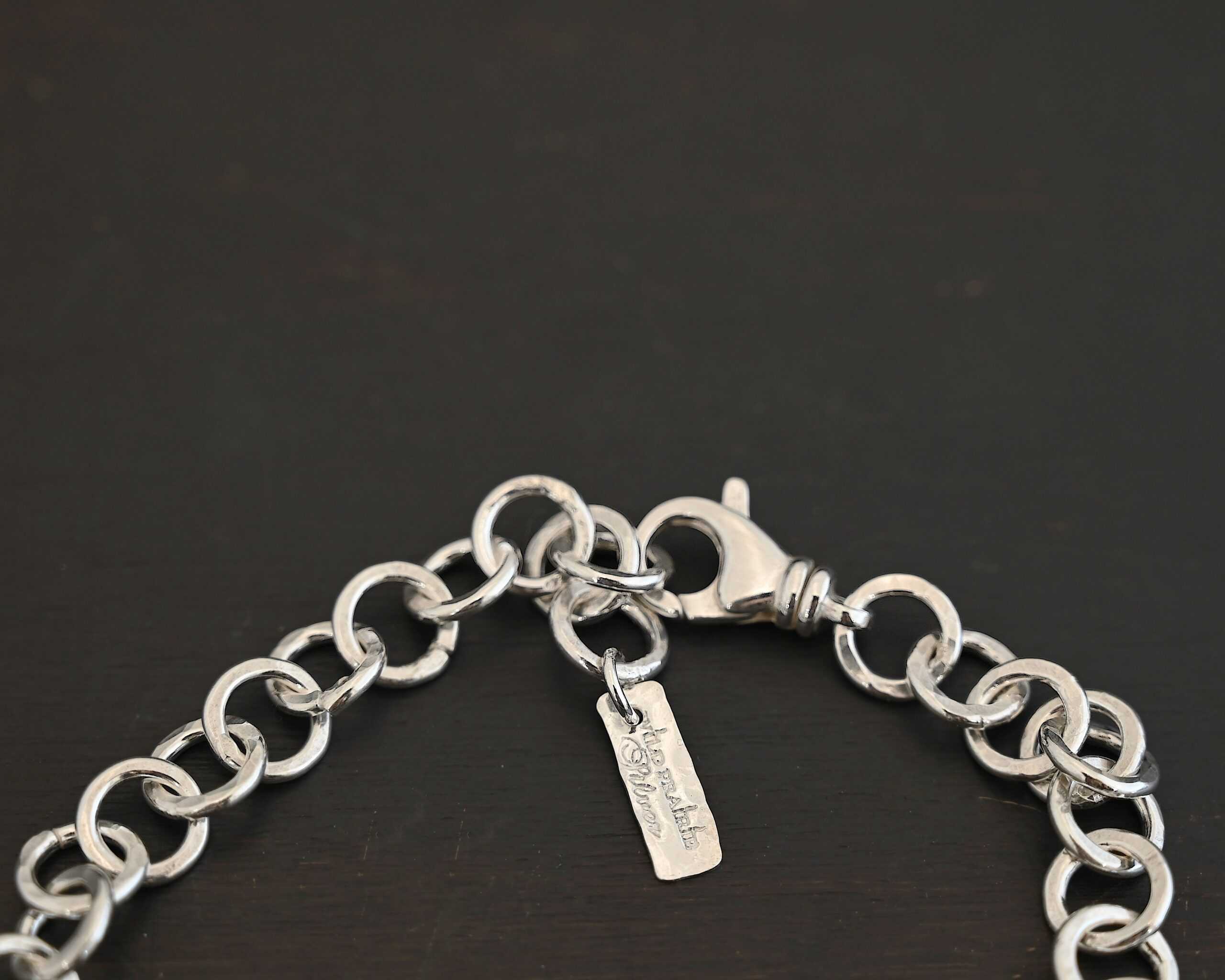 Close up of the "Wyatt" Chain. Chain is handmade out of sterling silver and has thick circle links that are hammered and shiny. chain has a lobster claw clasp and a rectangle tag that says "Wild Prairie Silver" on it. Chain is laying against a dark background.