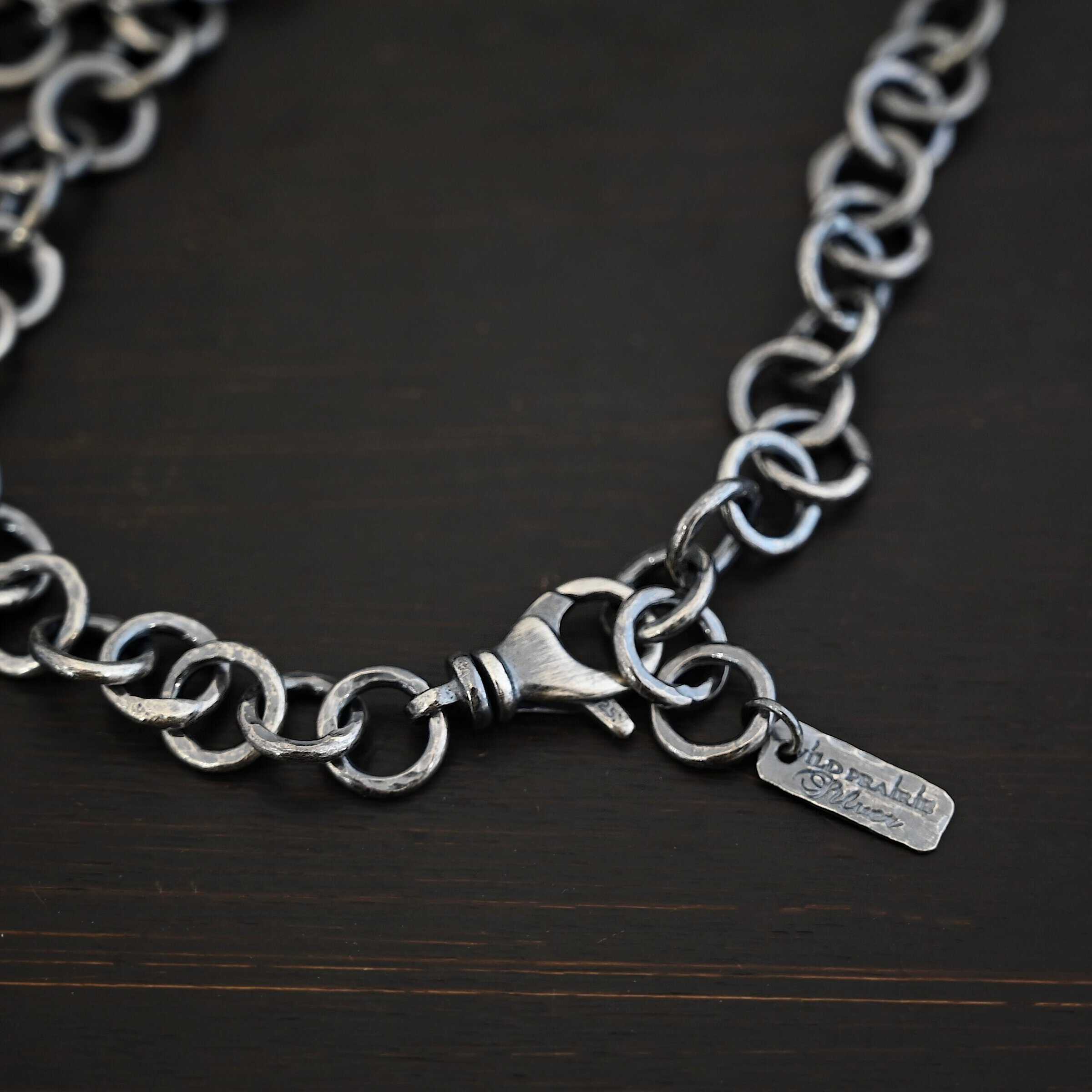 Close up of the "Wyatt" Chain. Chain is handmade out of sterling silver and has thick circle links that are hammered and oxidized. chain has a lobster claw clasp and a rectangle tag that says "Wild Prairie Silver" on it. Chain is laying against a dark background.