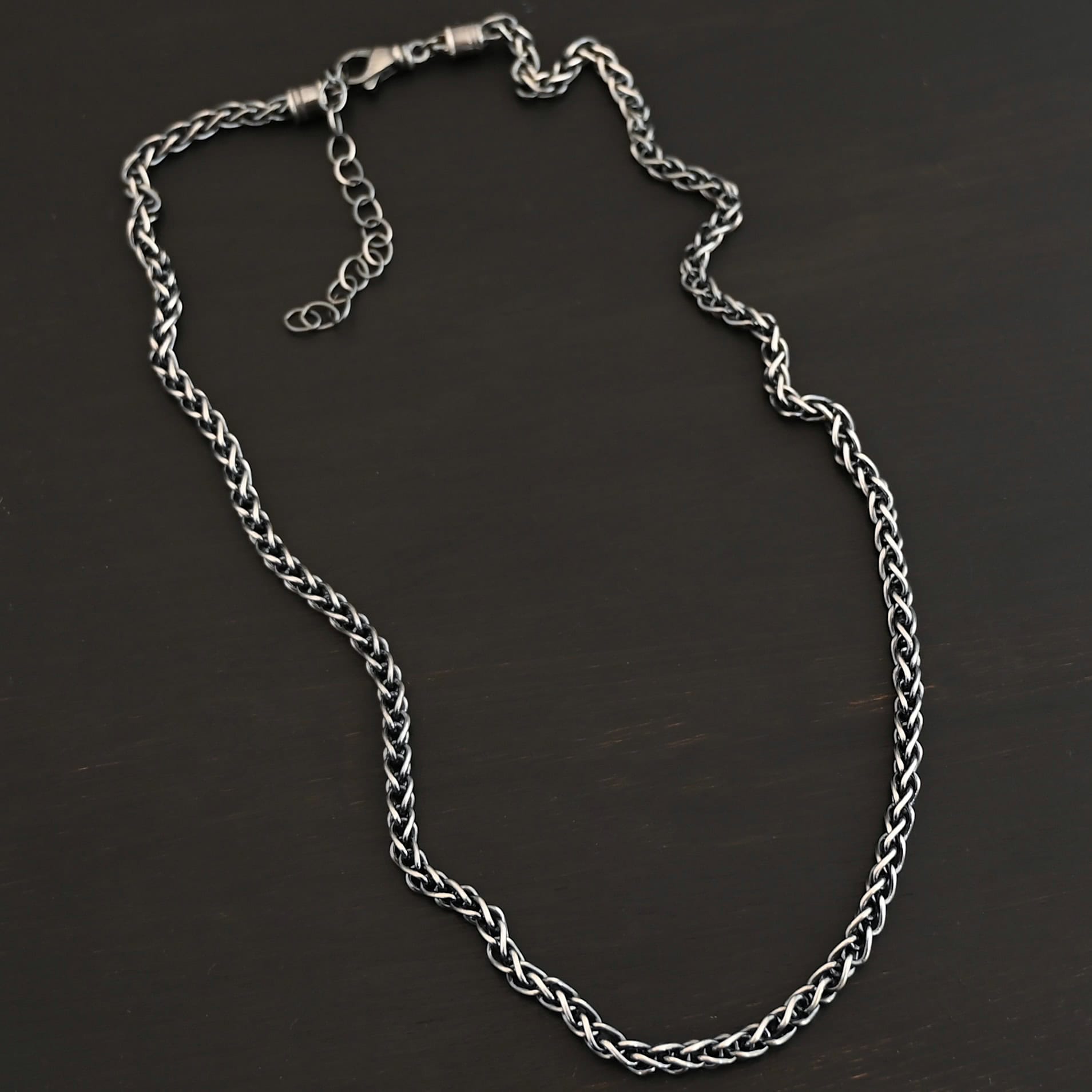 King Cobra Wheat Chain The "King Cobra" Wheat Chain. Chain is handmade out of sterling silver and woven together loosely. Each link is woven to another one and there are sterling silver end caps on each end of the chain. Chain has a lobster claw clasp and comes with a 3 inch extension. Chain is laying against a dark background. Chain is shown as oxidized.