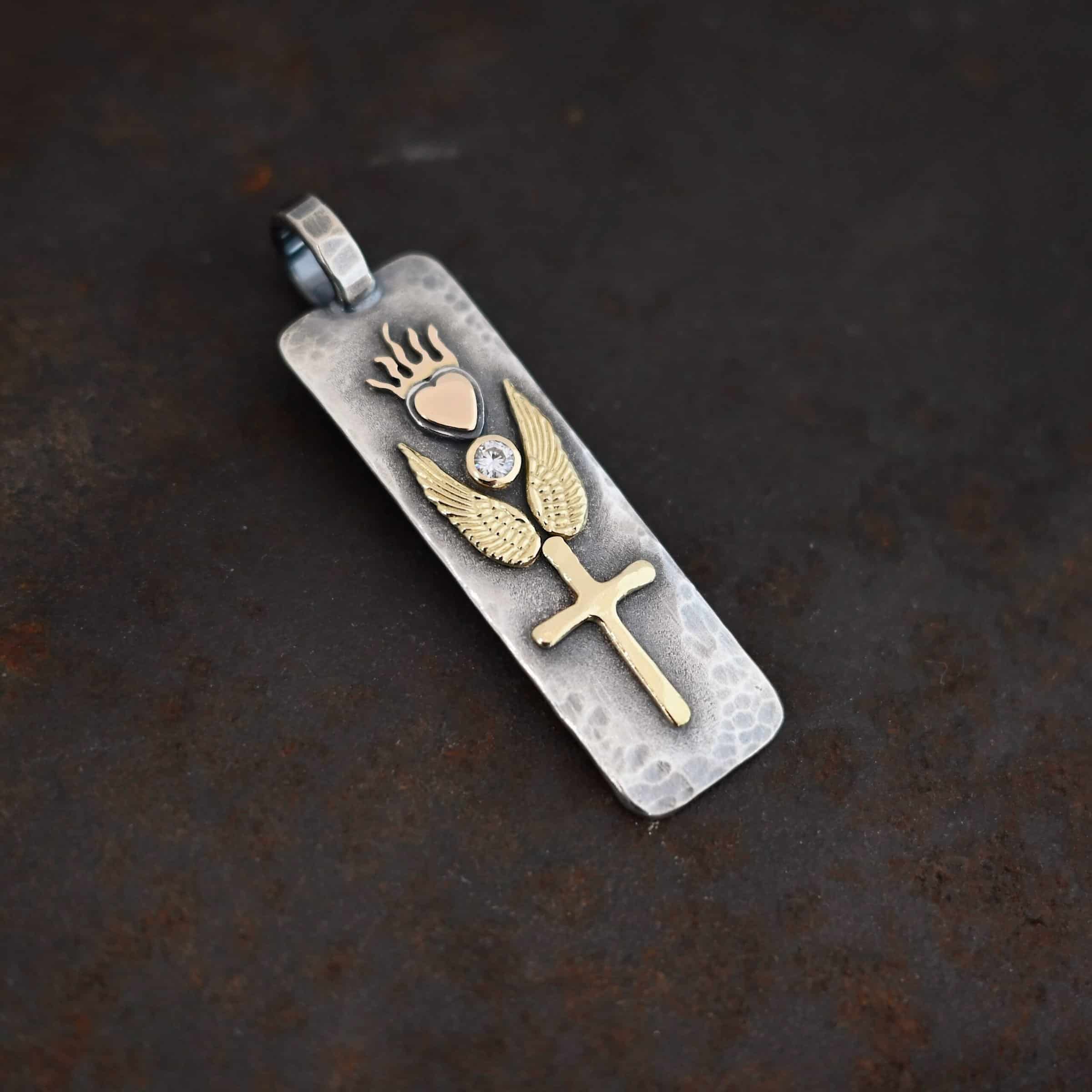 Protect Necklace Photo of the "Protect" Necklace. Necklace is made out of silver and gold. The pendant is a rectangle shaped silver tag that hangs vertically. It is large. On the top of the rectangle is a gold heart with gold flames dancing across the top of it. On the bottom, beneath the heart is a diamond that is set in gold. On each side of the diamond is a gold angel wing. Beneath the gold angel wings in the center of them and on the bottom of the rectangle pendant is a gold cross. There is a thick jump ring soldered to the top of the pendant. Necklace is laying against a dark background.