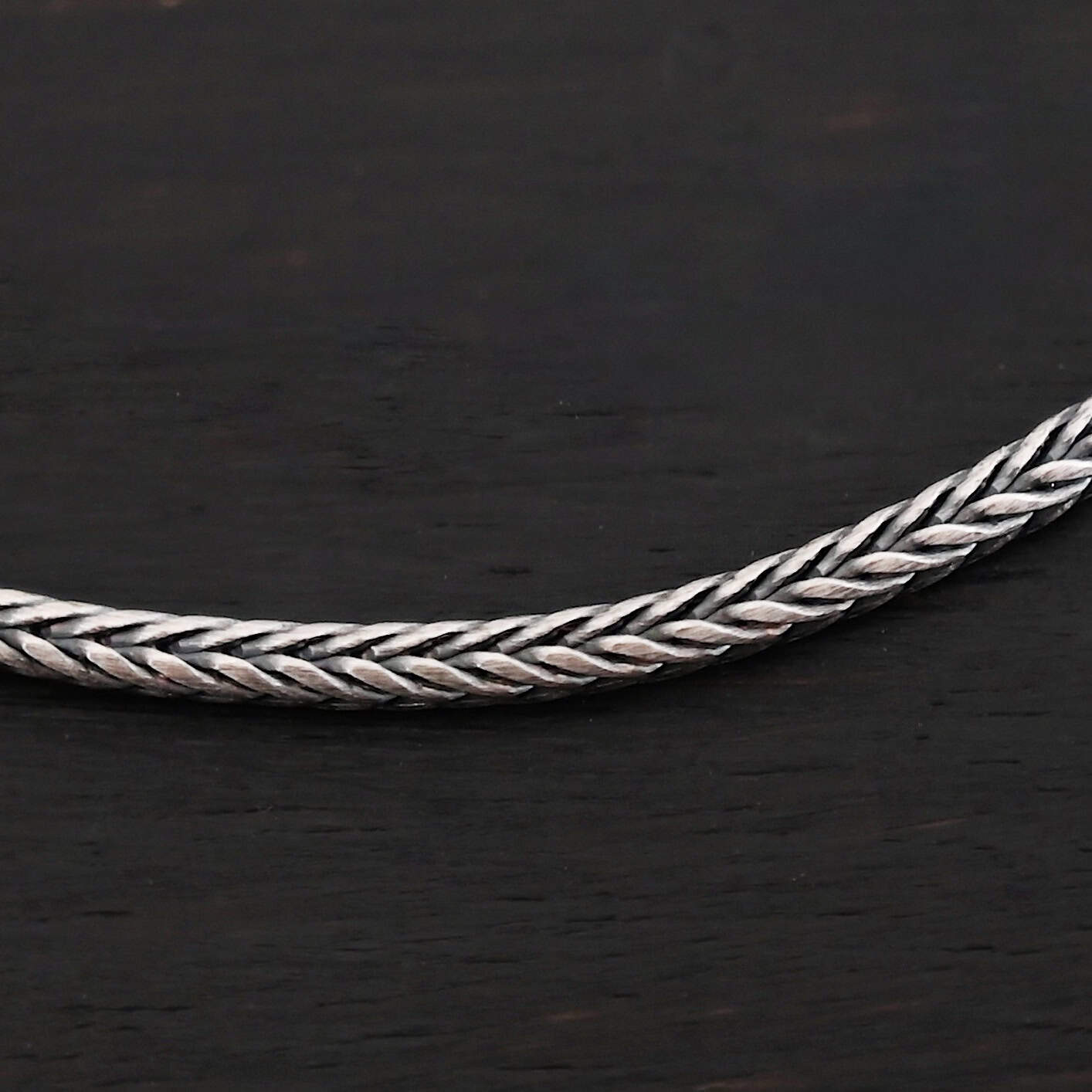 "Snake Foxtail" Chain. Chain is handmade out of sterling silver and woven together extremely tightly. Chain is oxidized. Chain is laying against a dark background.