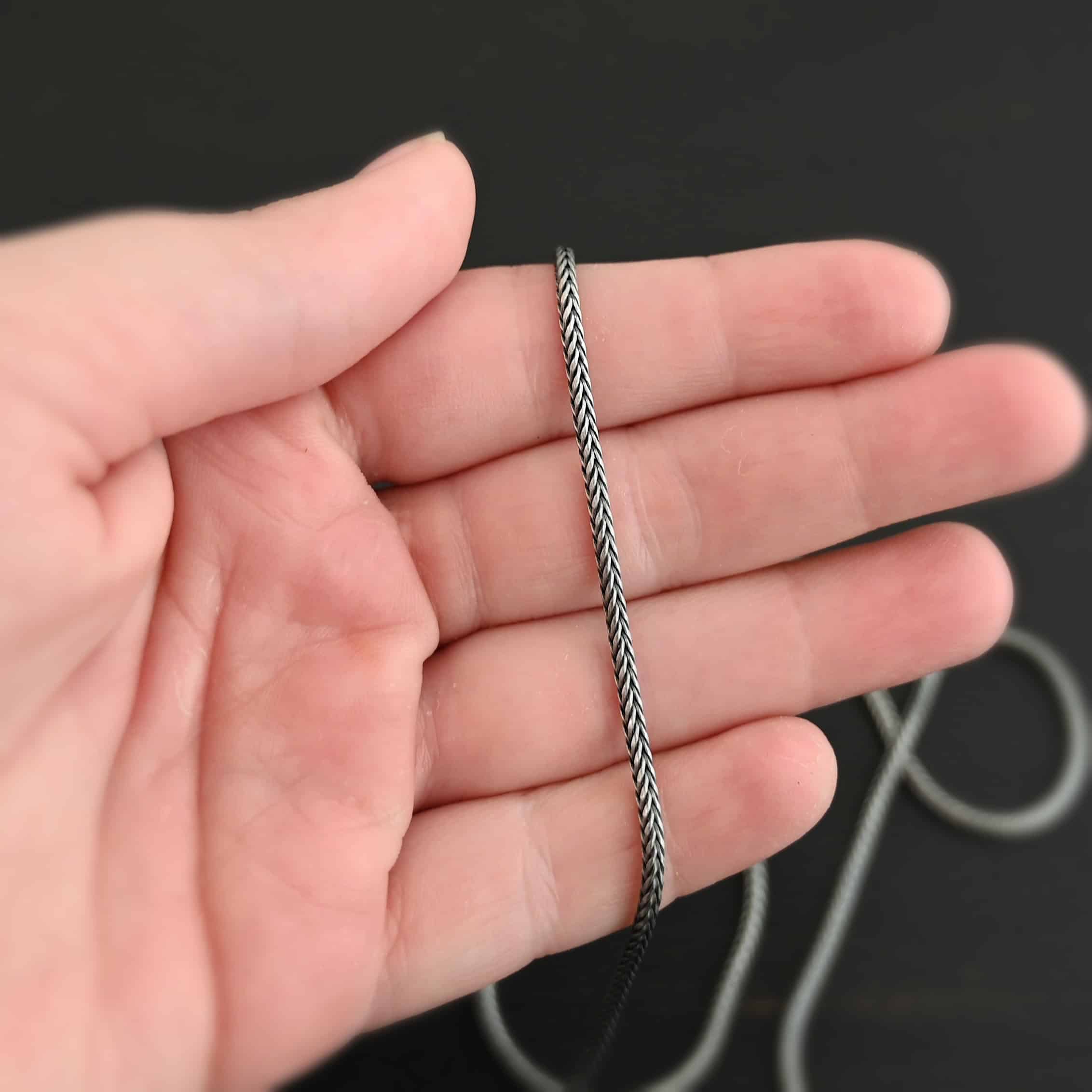 Oxidized sterling silver Snake Foxtail Chain that is woven together tightly to form the chain laying in a hand that is resting against a dark background