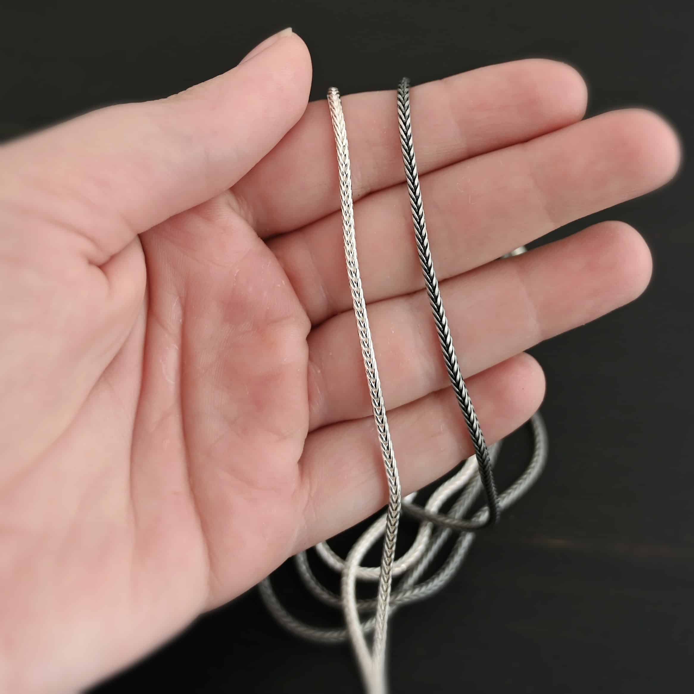 "Snake Foxtail" Chain. Chain is handmade out of sterling silver and woven together extremely tightly. Chain is shiny and oxidized. Chain is laying on a hand that is resting against a dark background.