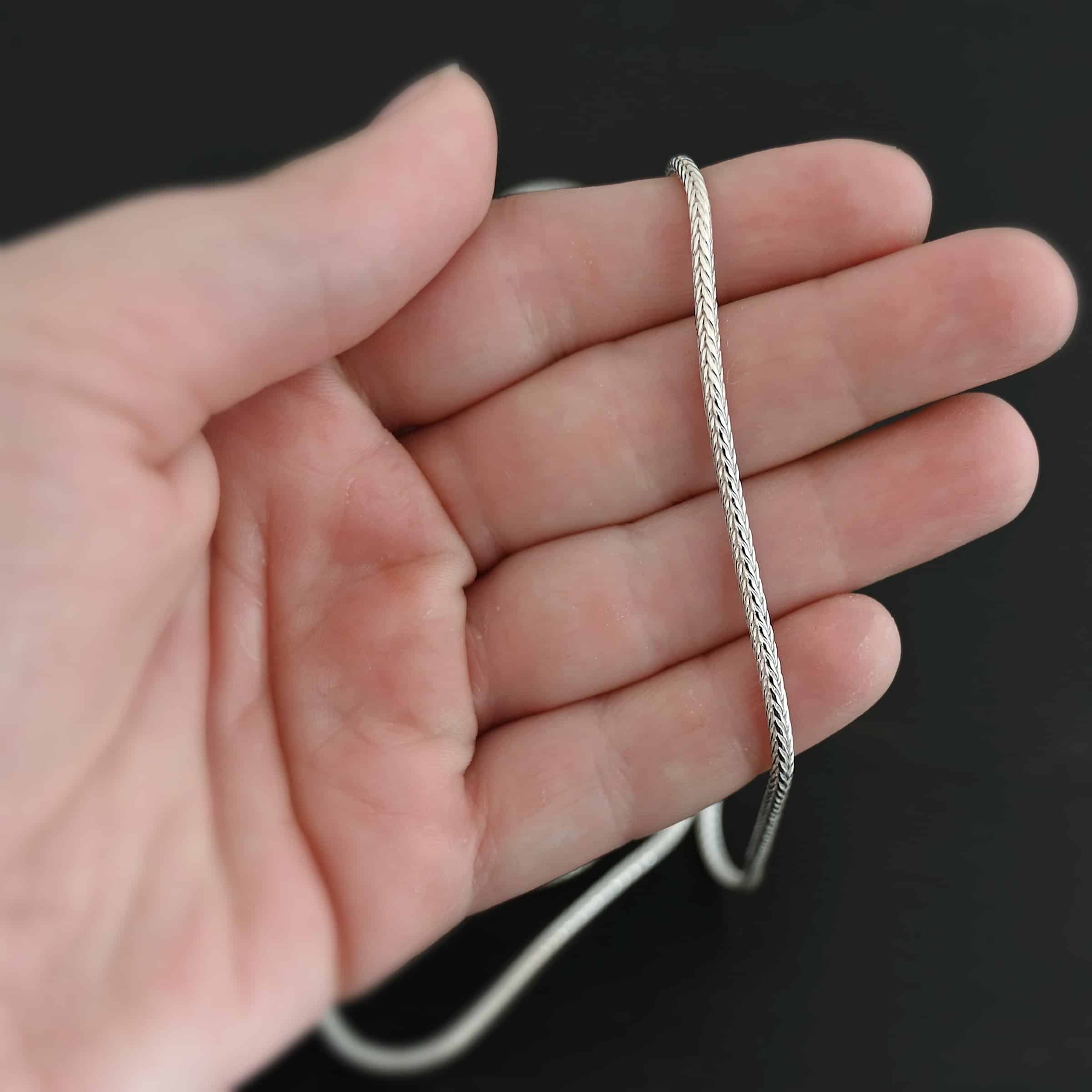 "Snake Foxtail" Chain. Chain is handmade out of sterling silver and woven together extremely tightly. Chain is shiny. Chain is laying on a hand that is resting against a dark background.
