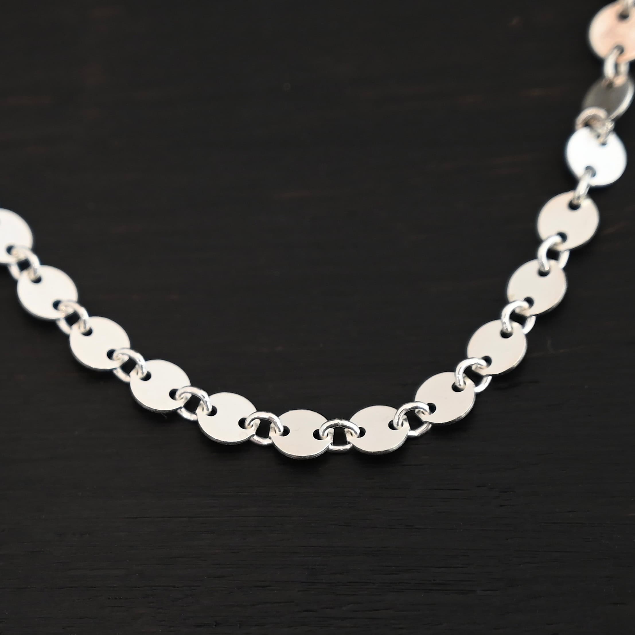 Sterling Silver Gypsy Coin Chain with circular disks that have two holes in each side of the disk that are connected by jump rings for the chain laying against a dark background.
