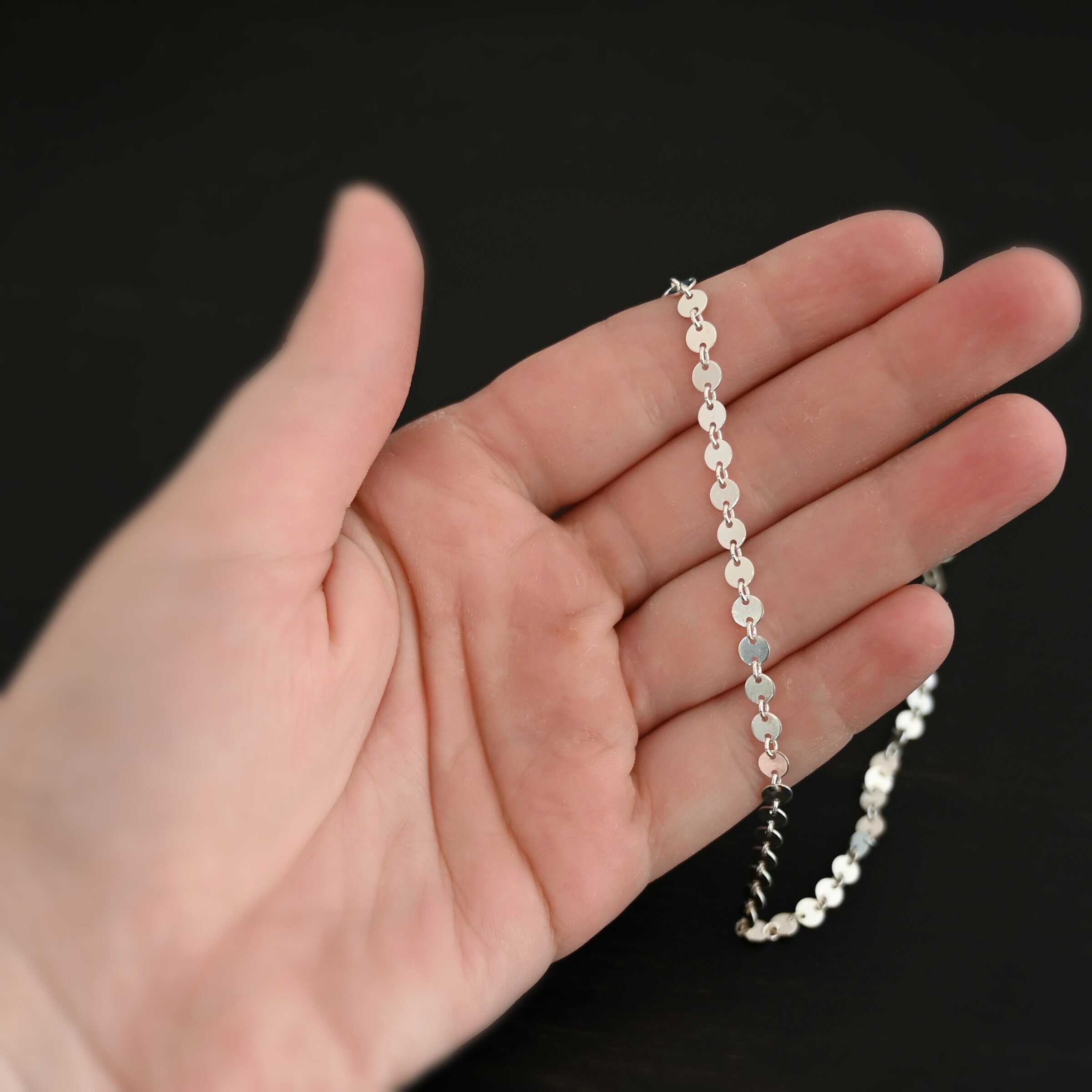 Sterling Silver Gypsy Coin Chain with circular disks for the chain laying on a hand that is resting against a dark background