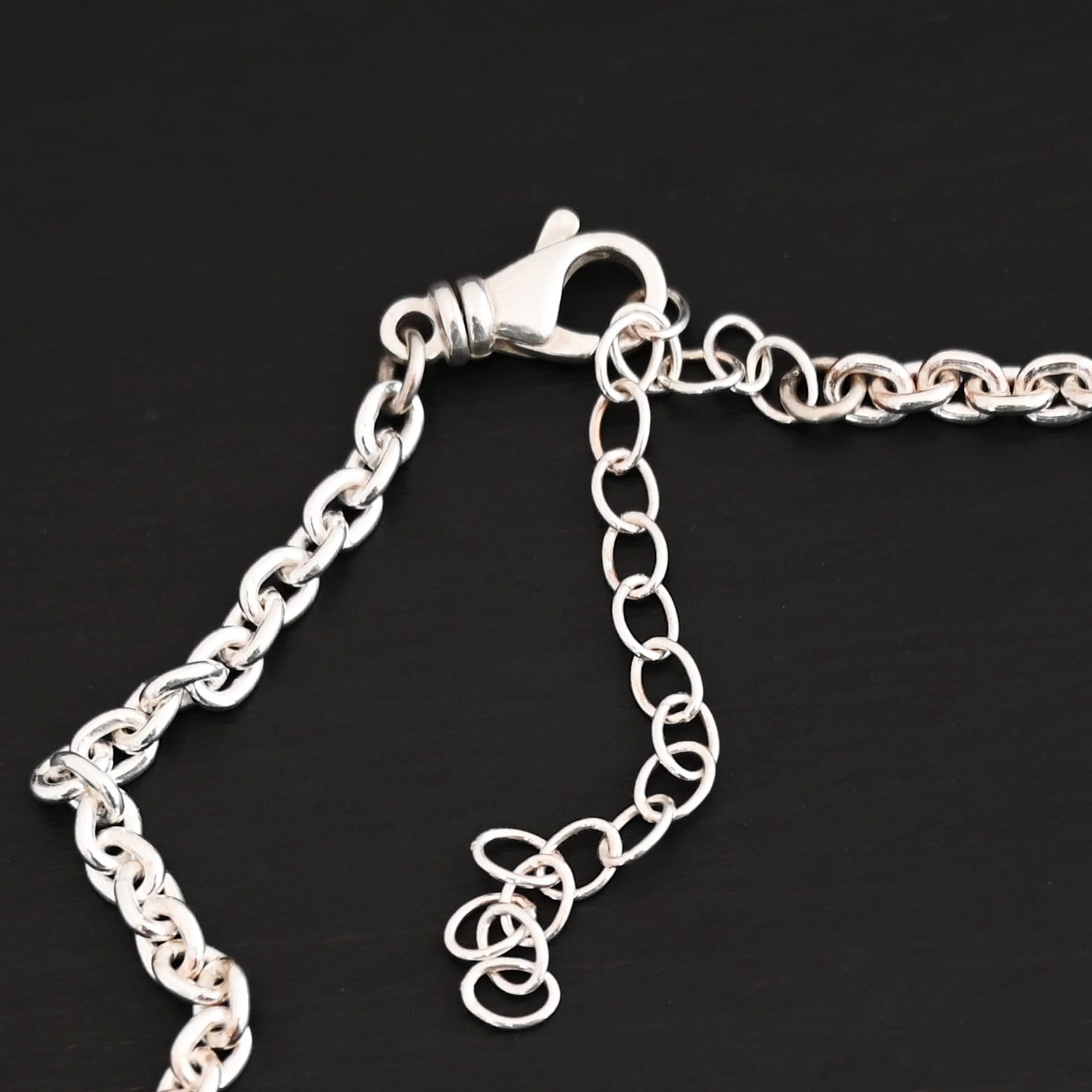 Joan Chain Shiny Close up of a shiny lobster claw clasp that is on a Shiny sterling silver Joan Chain with oval links that are 6x5mmx1.2mm thick that is laying on a dark background.