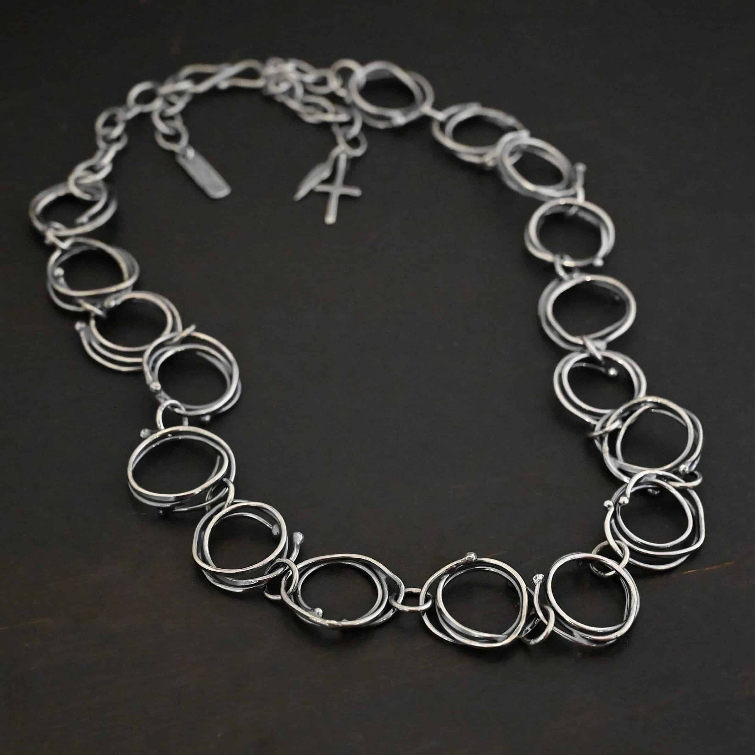 Photo of the "Organic Branch" Necklace. Made out of sterling silver. Necklace is made out of large circle links that are layered on top of one another unevenly so that each circle does not line up and you can tell there are multiple layers. There are silver dots scattered around the circles are well. Each circle is connected with a large jump ring. Necklace is laying against a black background. There is a cross and angel wing charm, as well as a tag on the chain in the background.