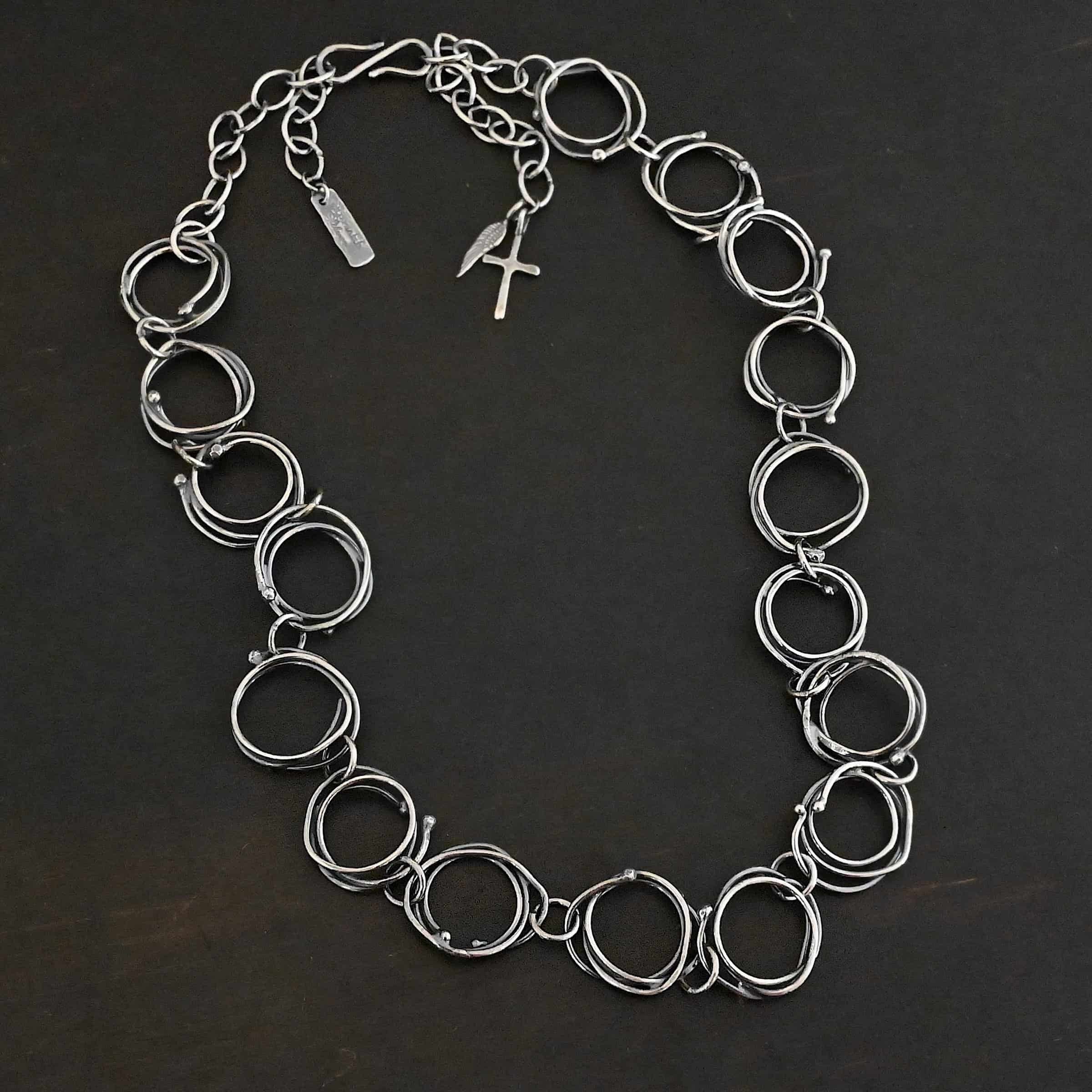 Photo of the "Organic Branch" Necklace. Made out of sterling silver. Necklace is made out of large circle links that are layered on top of one another unevenly so that each circle does not line up and you can tell there are multiple layers. There are silver dots scattered around the circles are well. Each circle is connected with a large jump ring. Necklace is laying against a black background.