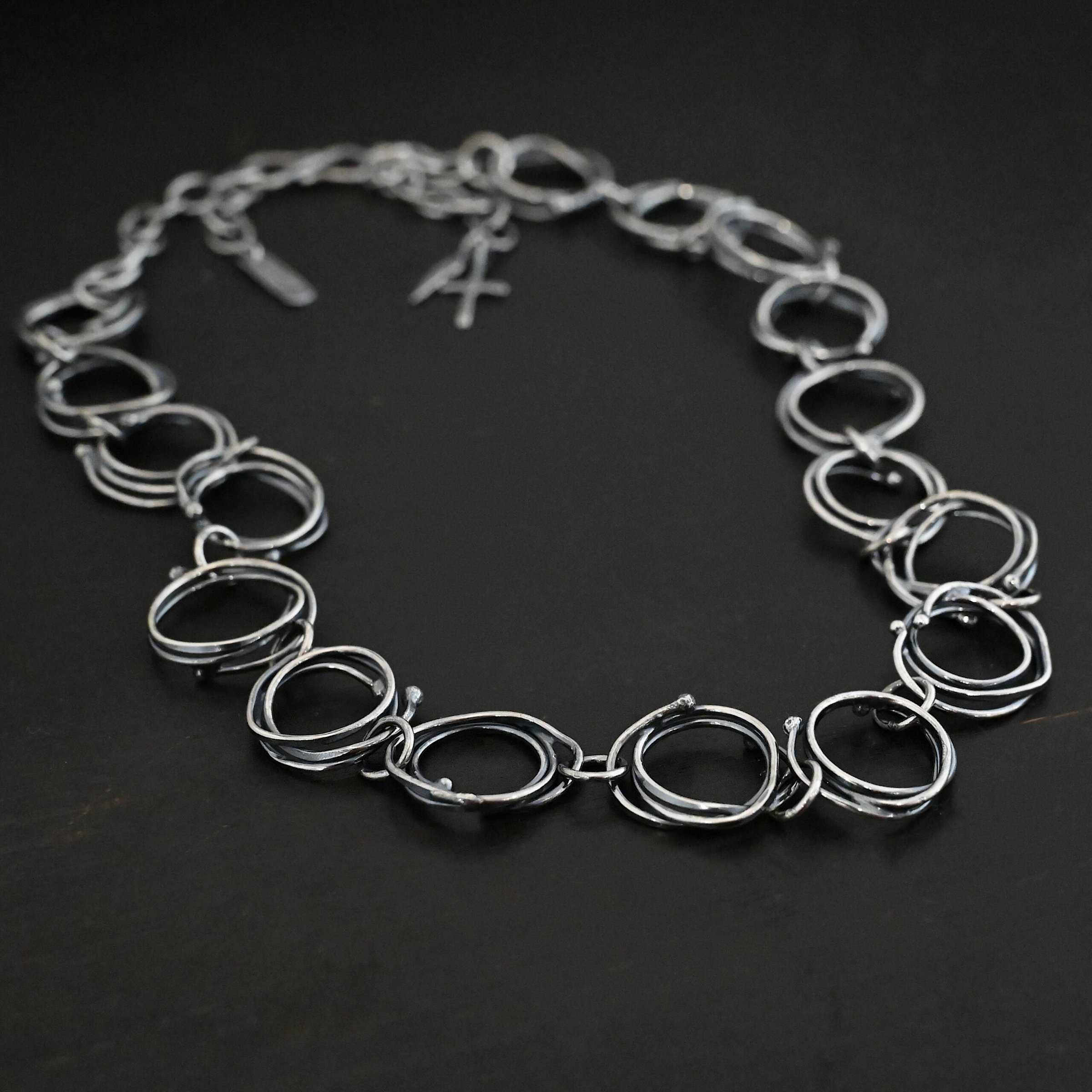 Photo of the "Organic Branch" Necklace. Made out of sterling silver. Necklace is made out of large circle links that are layered on top of one another unevenly so that each circle does not line up and you can tell there are multiple layers. There are silver dots scattered around the circles are well. Each circle is connected with a large jump ring. Necklace is laying against a black background.