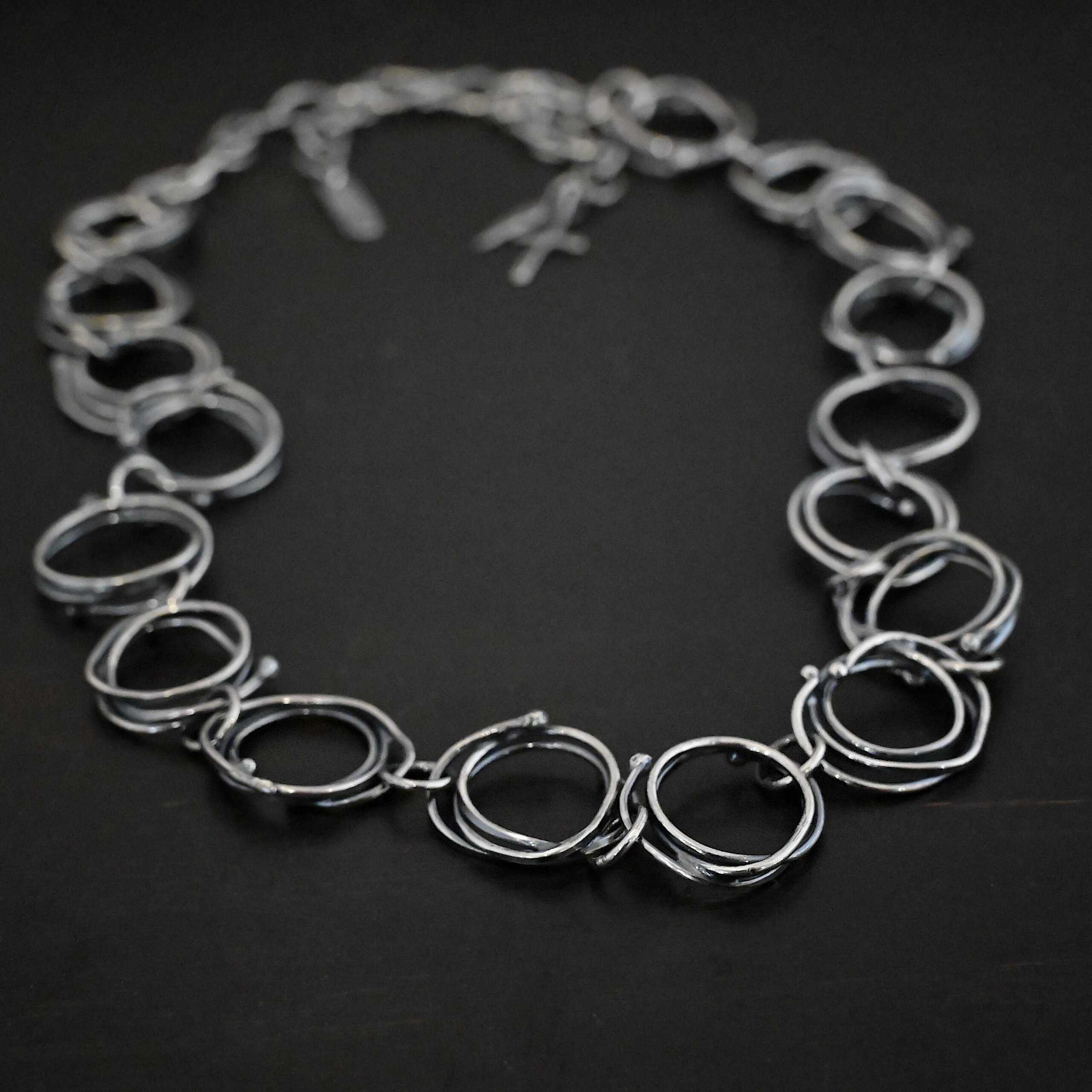 Photo of the "Organic Branch" Necklace. Made out of sterling silver. Necklace is made out of large circle links that are layered on top of one another unevenly so that each circle does not line up and you can tell there are multiple layers. There are silver dots scattered around the circles are well. Each circle is connected with a large jump ring. Necklace is laying against a black background.