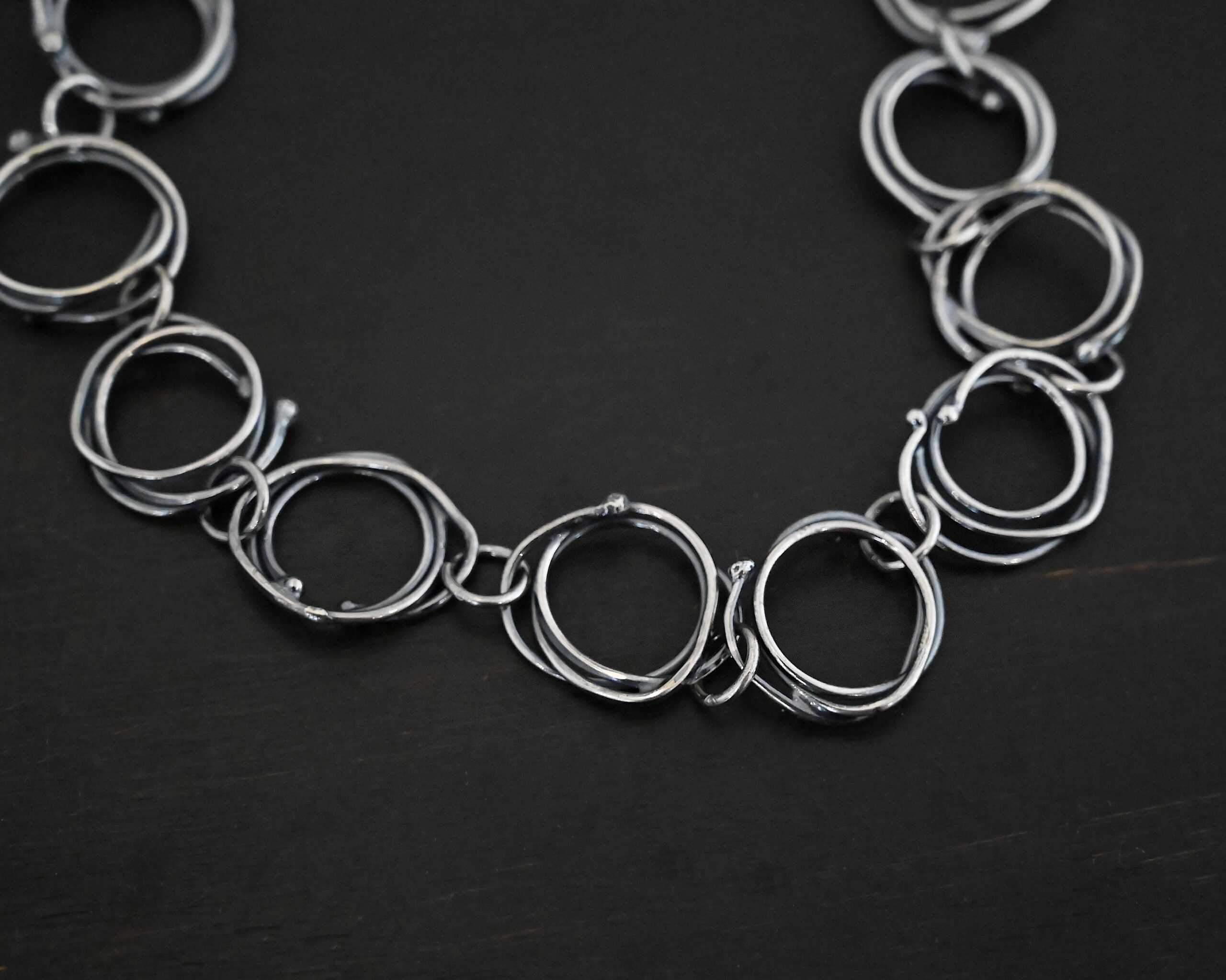 Photo of the "Organic Branch" Necklace. Made out of sterling silver. Necklace is made out of large circle links that are layered on top of one another unevenly so that each circle does not line up and you can tell there are multiple layers. There are silver dots scattered around the circles are well. Each circle is connected with a large jump ring. Necklace is laying against a black background.