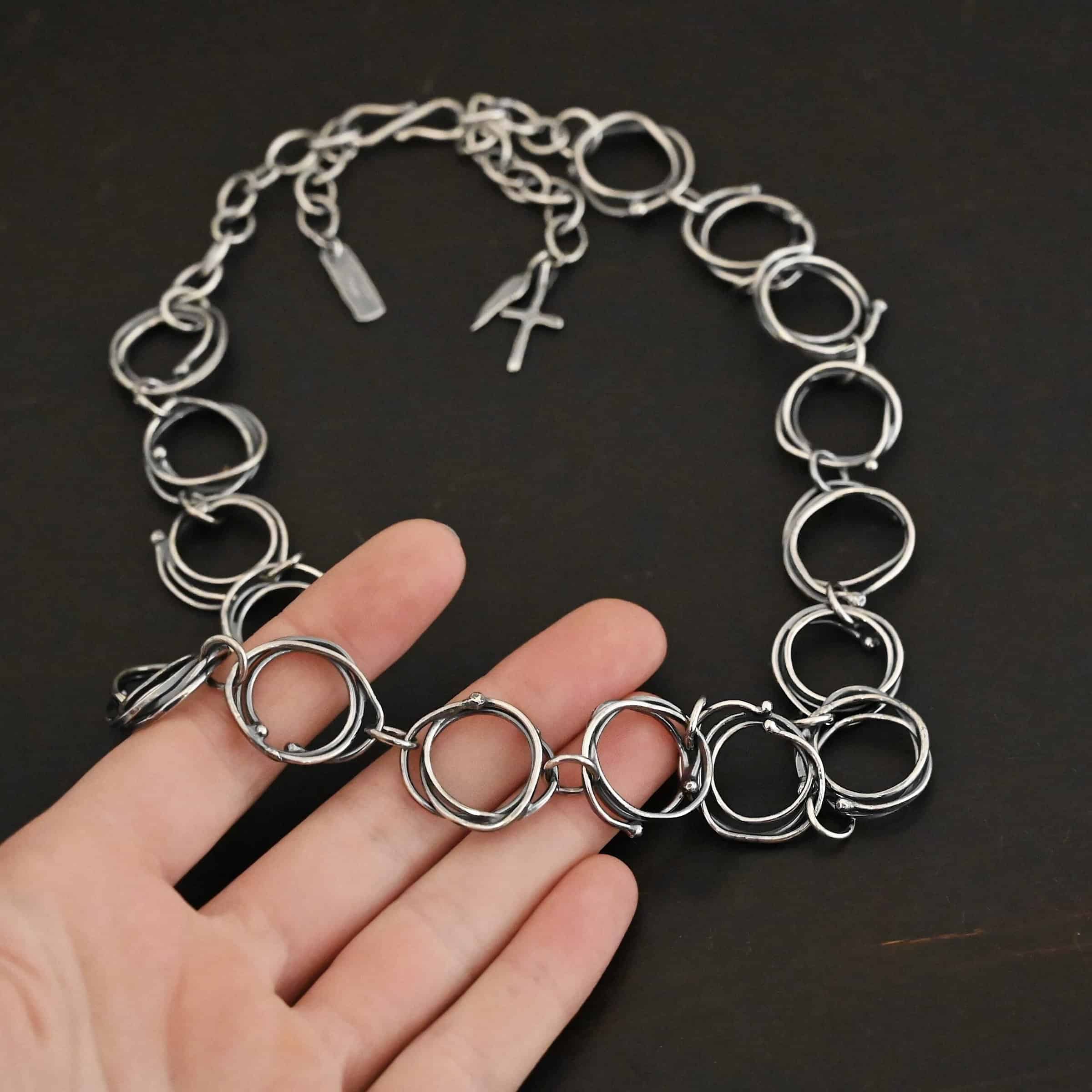 Photo of the "Organic Branch" Necklace. Made out of sterling silver. Necklace is made out of large circle links that are layered on top of one another unevenly so that each circle does not line up and you can tell there are multiple layers. There are silver dots scattered around the circles are well. Each circle is connected with a large jump ring. Necklace is laying against the fingertips of a hand to demonstrate size. There is a cross and angel wing charm, as well as a tag on the chain in the background.