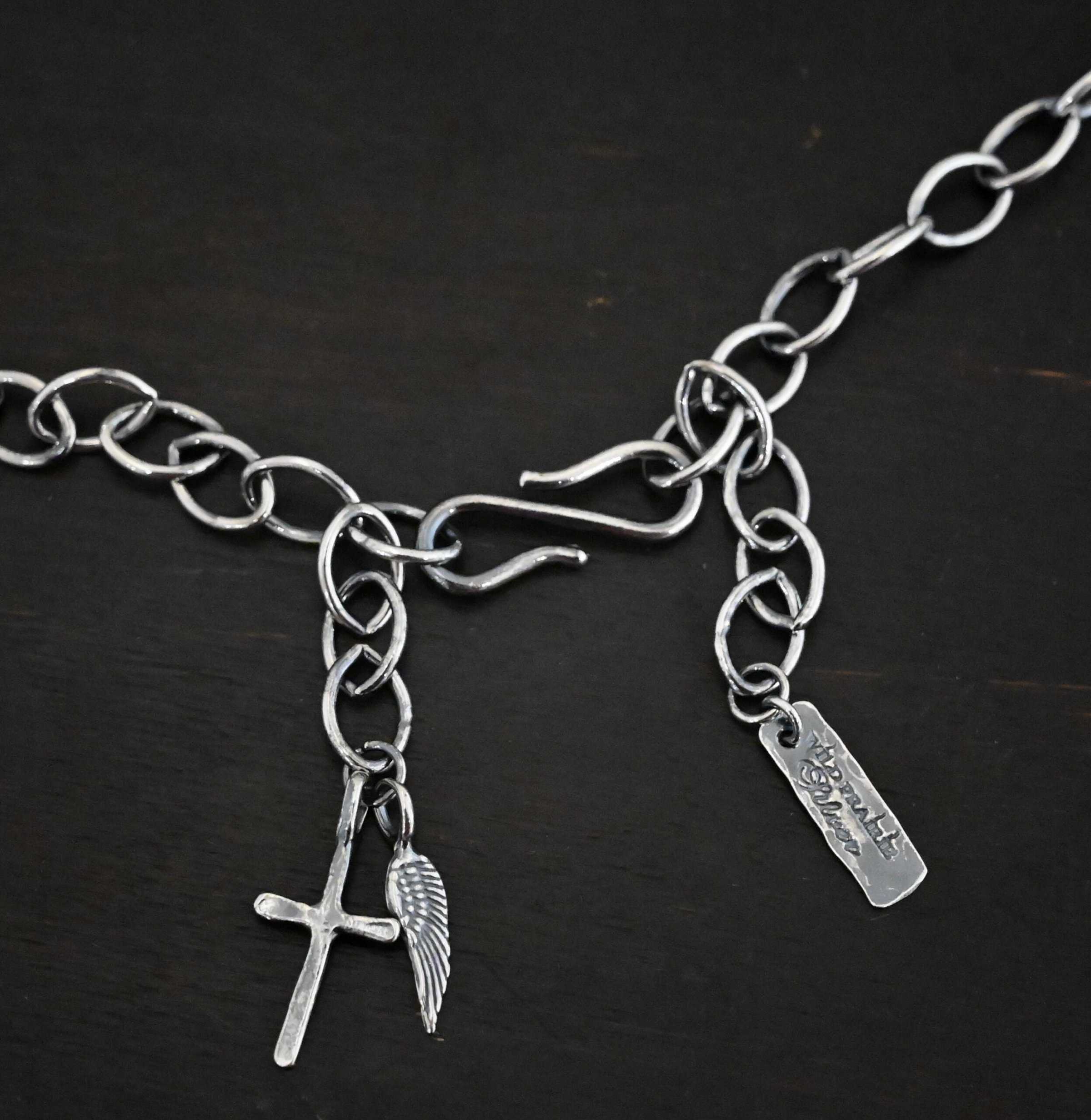 Chain of the "Organic Branch" Necklace. Made out of sterling silver. Chain has oval links. There are two sides of the chain that meet at a figure eight hook clasp in the center. On the left side of the chain at the end is a cross and angel wing charm. On the right side at the end of the chain is a silver tag that says "Wild Prairie Silver." Chain is laying against a dark background.