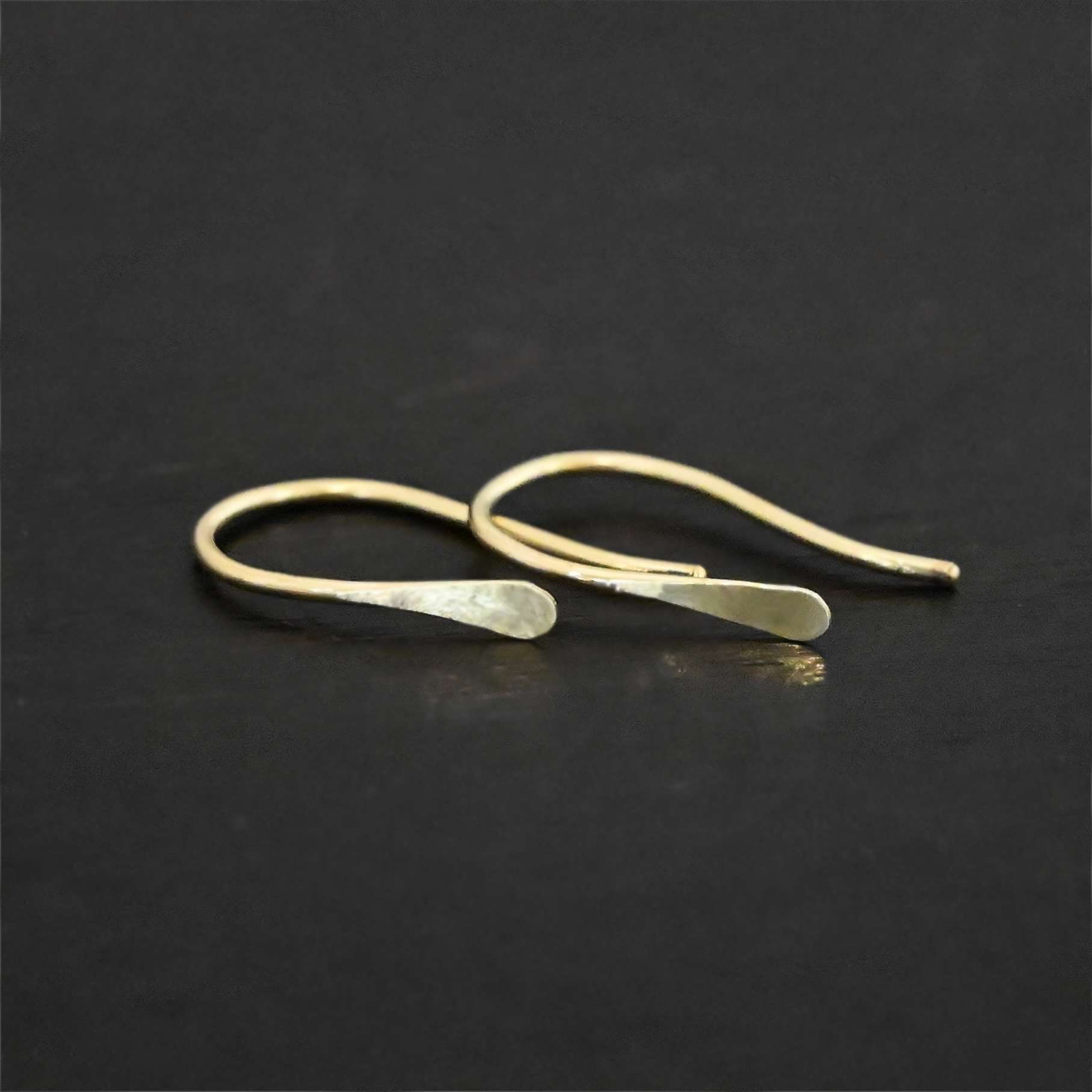 18k Gold Roadies Close up of the "Roadie" Earrings. Made out of 18K gold. Earrings are small and thin. Earrings are a French hook ear wire with a hammered end that looks like a canoe paddle. Earrings are laying against a dark background.