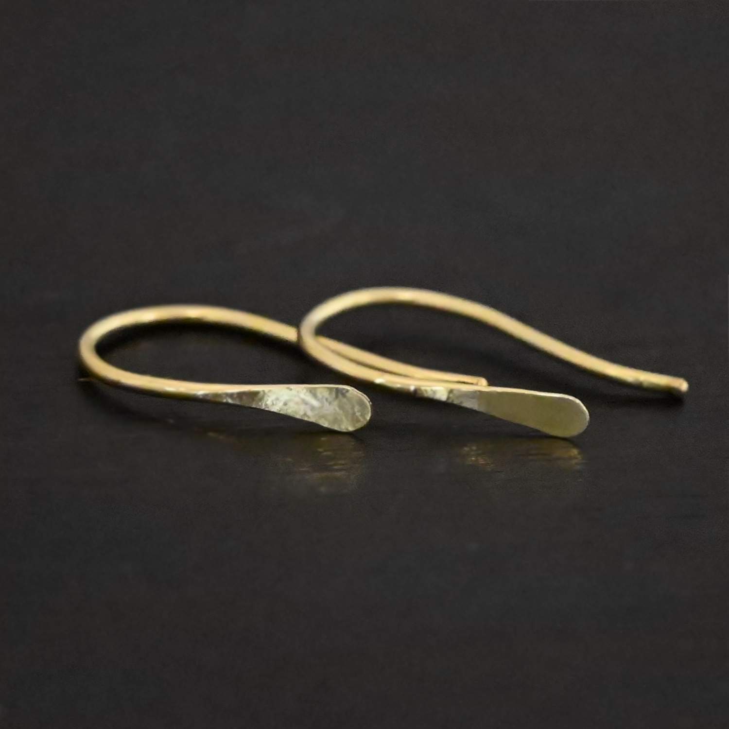 18k Gold Roadies Close up of the "Roadie" Earrings. Made out of 18K gold. Earrings are small and thin. Earrings are a French hook ear wire with a hammered end that looks like a canoe paddle. Earrings are laying against a dark background.