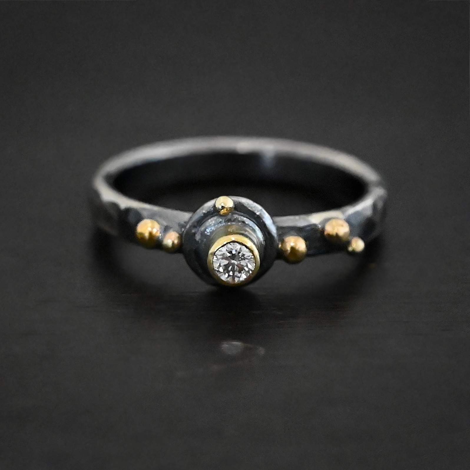 Prairie Princess Ring Close up of the "Prairie Princess" Ring. Made out of sterling silver, gold, and diamond. There is a small circular diamond for the center stone that is set in gold. The gold setting is set on two layers of sterling silver. Each layer is a flat circular disk. On the second disk at the top of it is a small gold dot. On the left side of the diamond on the ring band are two small gold dots. On the right side of the diamond on the band are three gold dots. Ring is sitting against a dark background.