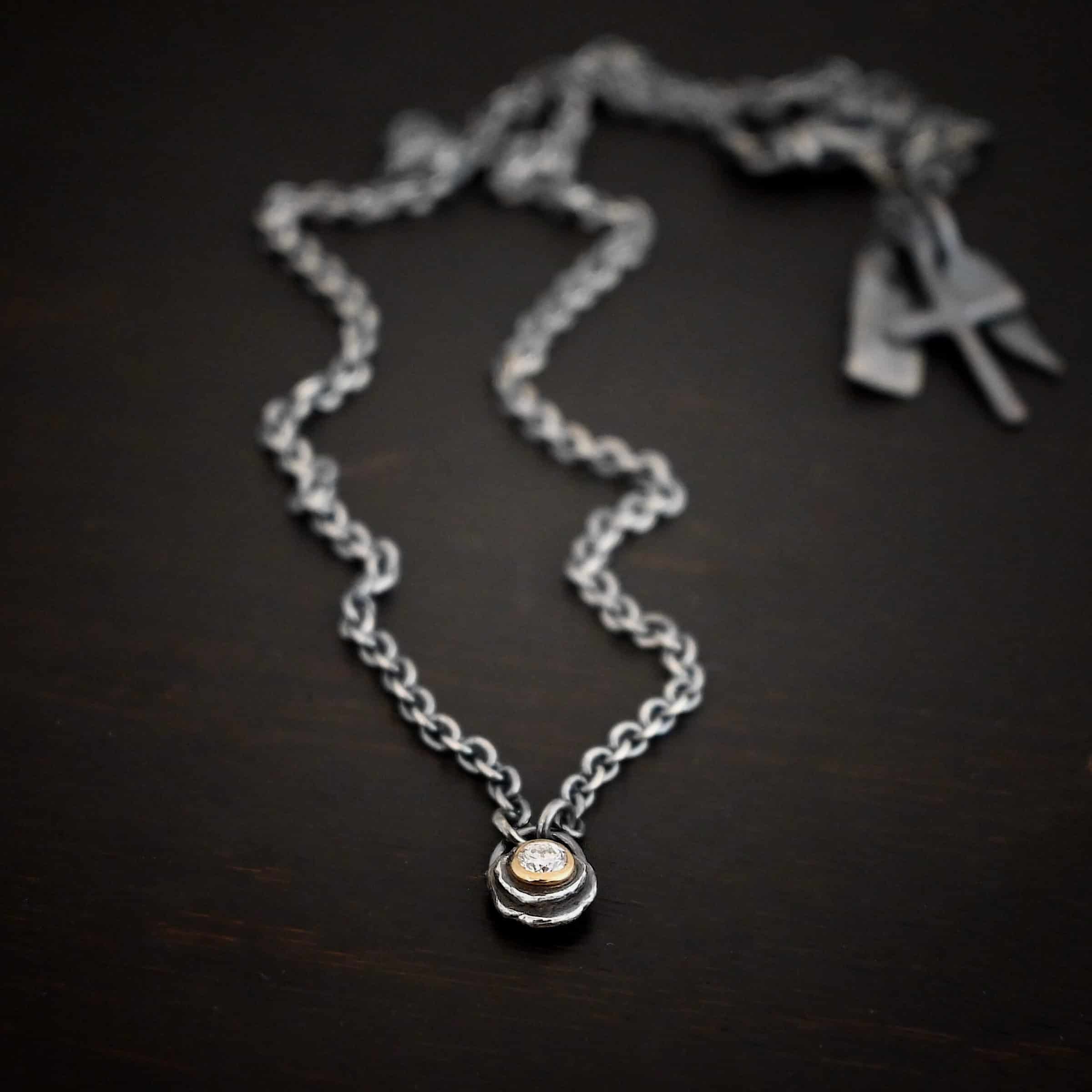 Audrey Hepburn Necklace Photo of the "Audrey Hepburn" Necklace. There is a small diamond in the center of a small circle pendant. The diamond is set in gold. There are two layers of sterling silver that are both hammered flat dots. One layer of silver is smaller, and sits on top of the bigger layer of silver. The gold is on the very top of the pendant. There is a small loop soldered to the back of the necklace in which two silver jump rings hang from. Each end of the chain hangs on the jump rings. Necklace is lying against a dark background.