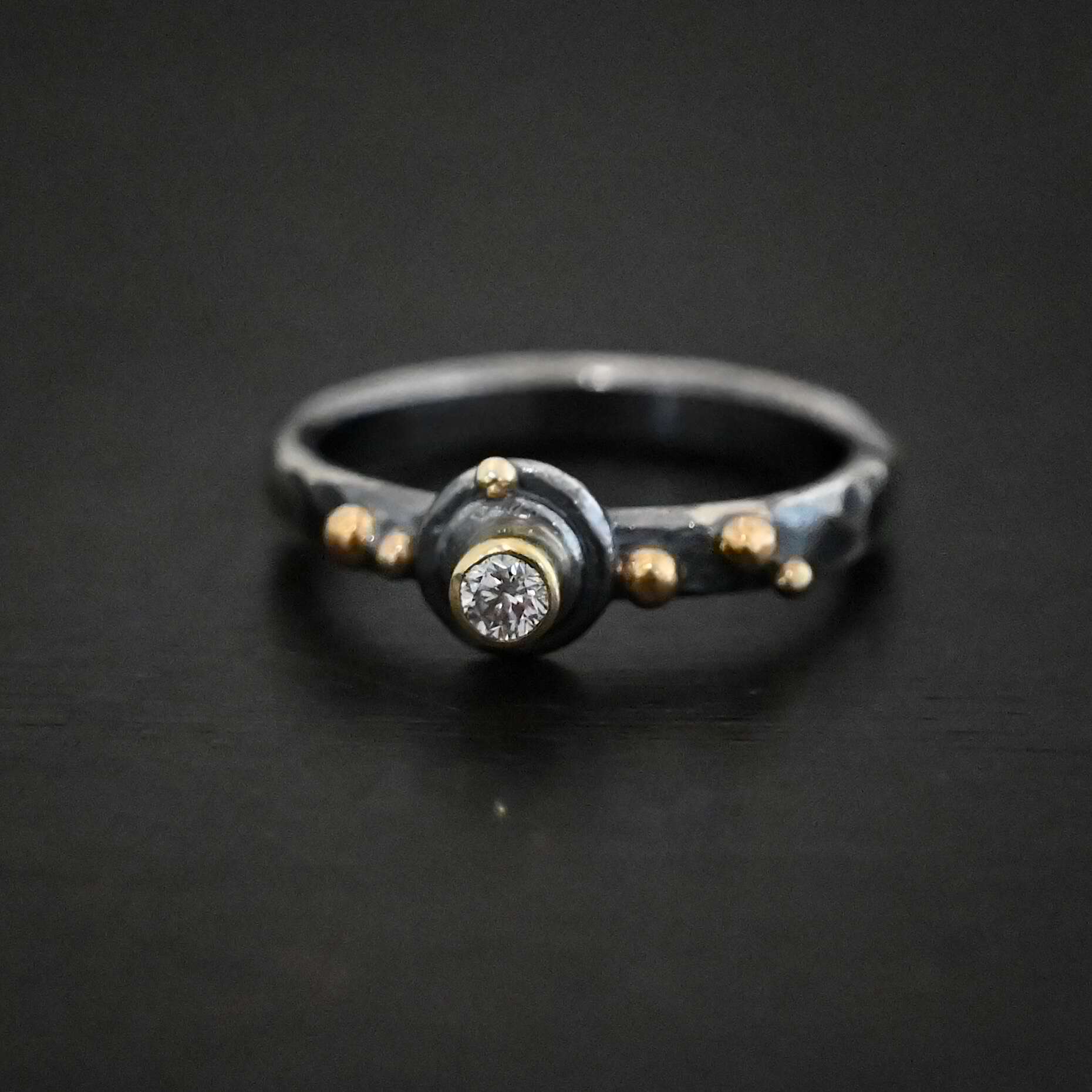 Prairie Princess Ring Close up of the "Prairie Princess" Ring. Made out of sterling silver, gold, and diamond. There is a small circular diamond for the center stone that is set in gold. The gold setting is set on two layers of sterling silver. Each layer is a flat circular disk. On the second disk at the top of it is a small gold dot. On the left side of the diamond on the ring band are two small gold dots. On the right side of the diamond on the band are three gold dots. Ring is sitting against a dark background.