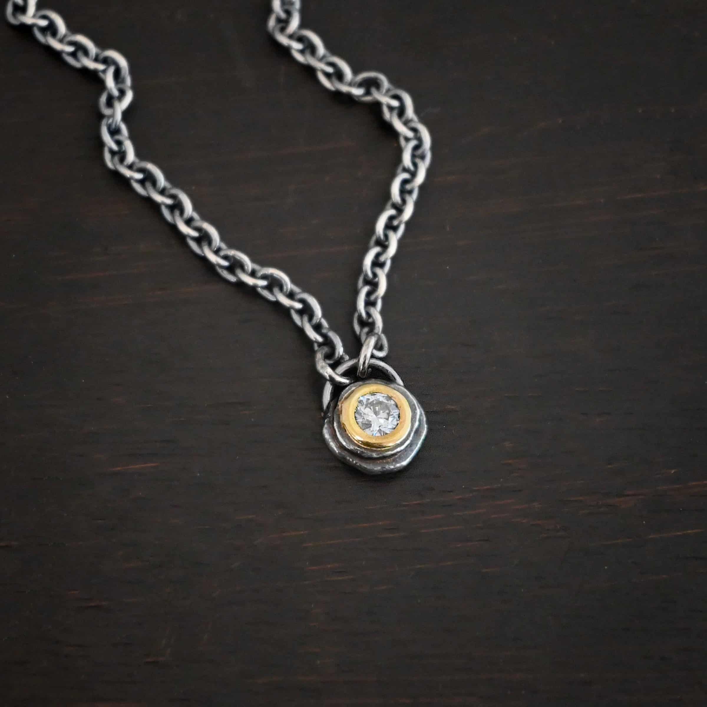 Audrey Hepburn Necklace Photo of the "Audrey Hepburn" Necklace. There is a small diamond in the center of a small circle pendant. The diamond is set in gold. There are two layers of sterling silver that are both hammered flat dots. One layer of silver is smaller, and sits on top of the bigger layer of silver. The gold is on the very top of the pendant. There is a small loop soldered to the back of the necklace in which two silver jump rings hang from. Each end of the chain hangs on the jump rings. Necklace is lying against a dark background.